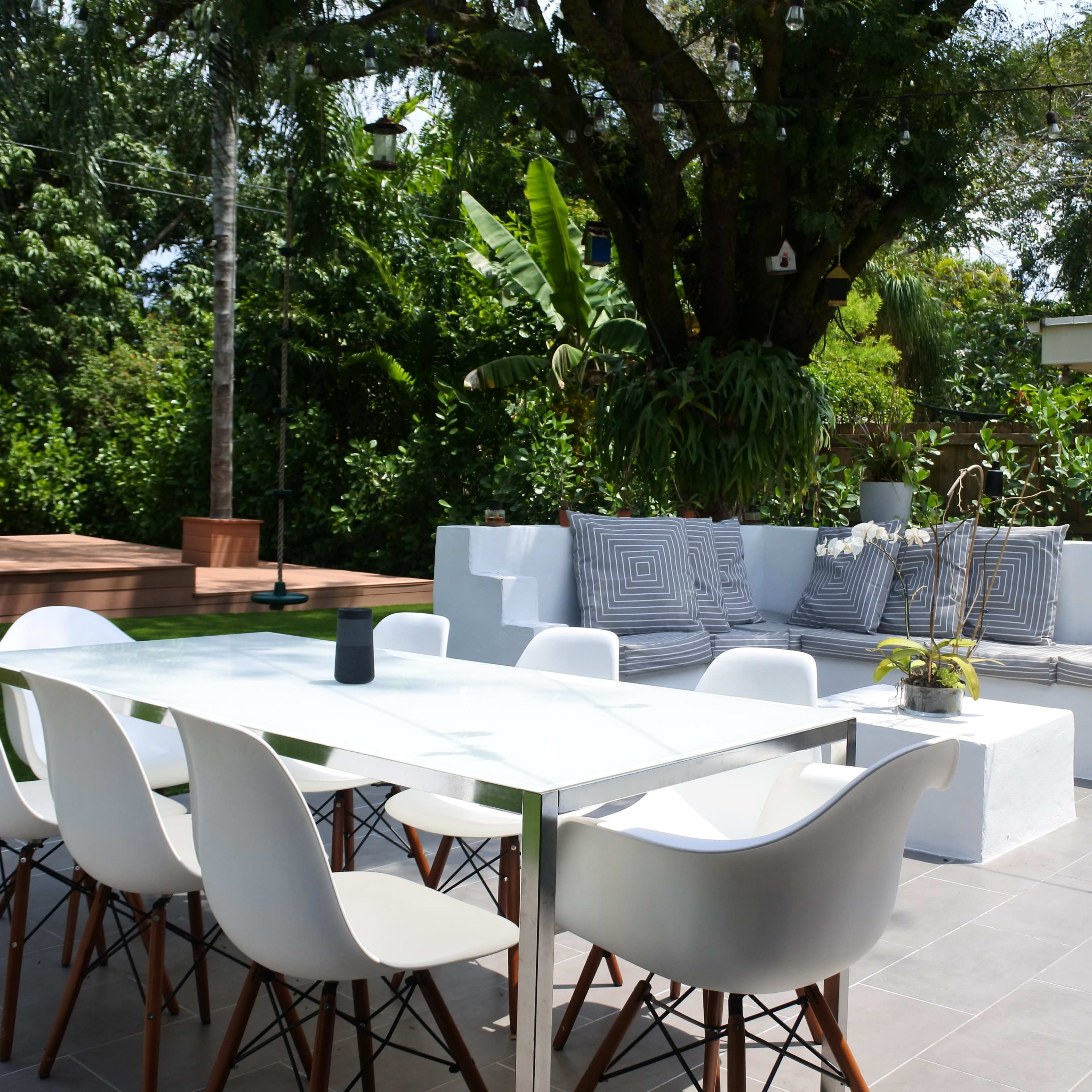 A modern outdoor dining area features a glass table surrounded by white chairs, with a plush seating arrangement nearby and a swimming pool in the background.