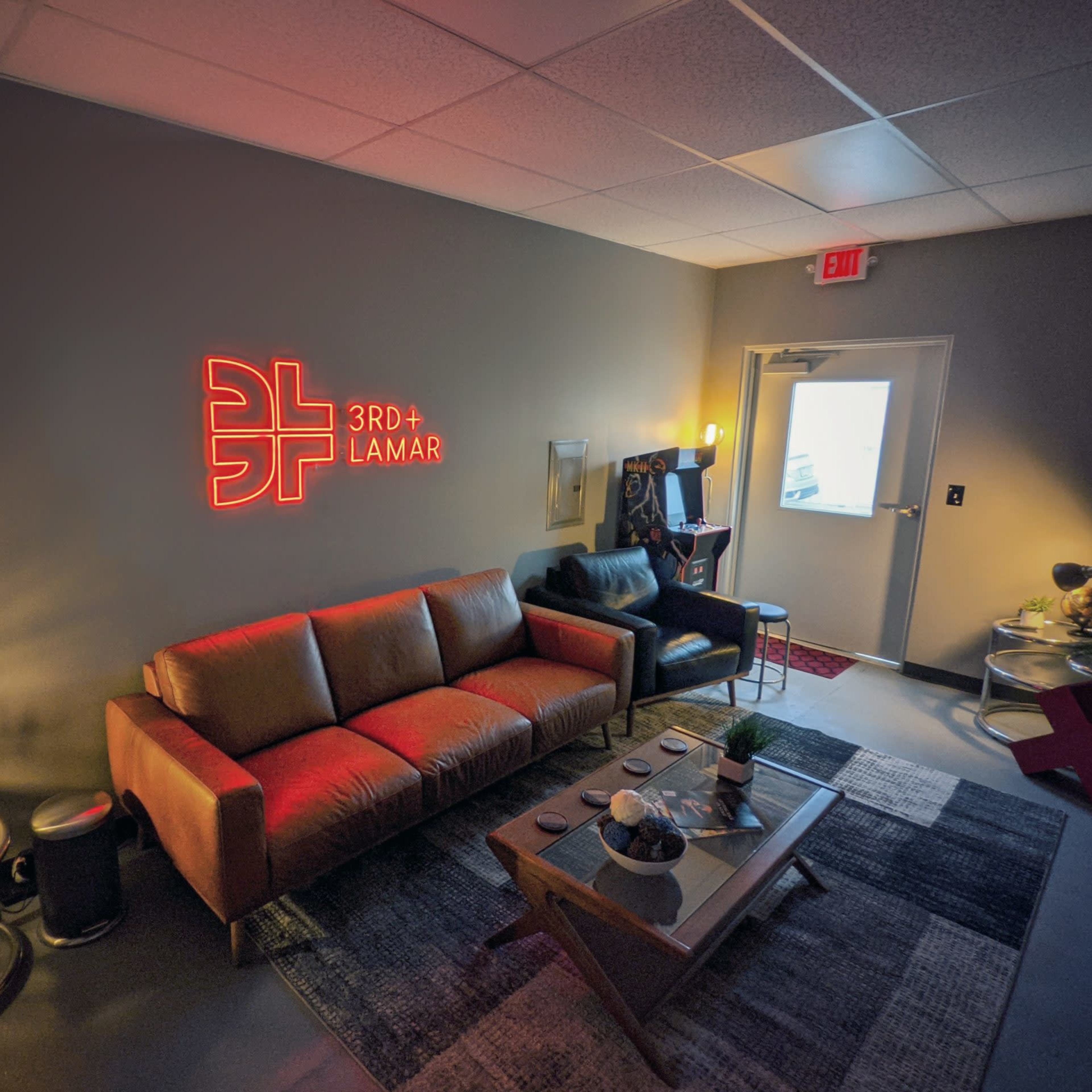 A modern lounge area with a glowing neon sign that reads "3RD + LAMAR," featuring a brown sofa, a black chair, and various decorative items on a table.