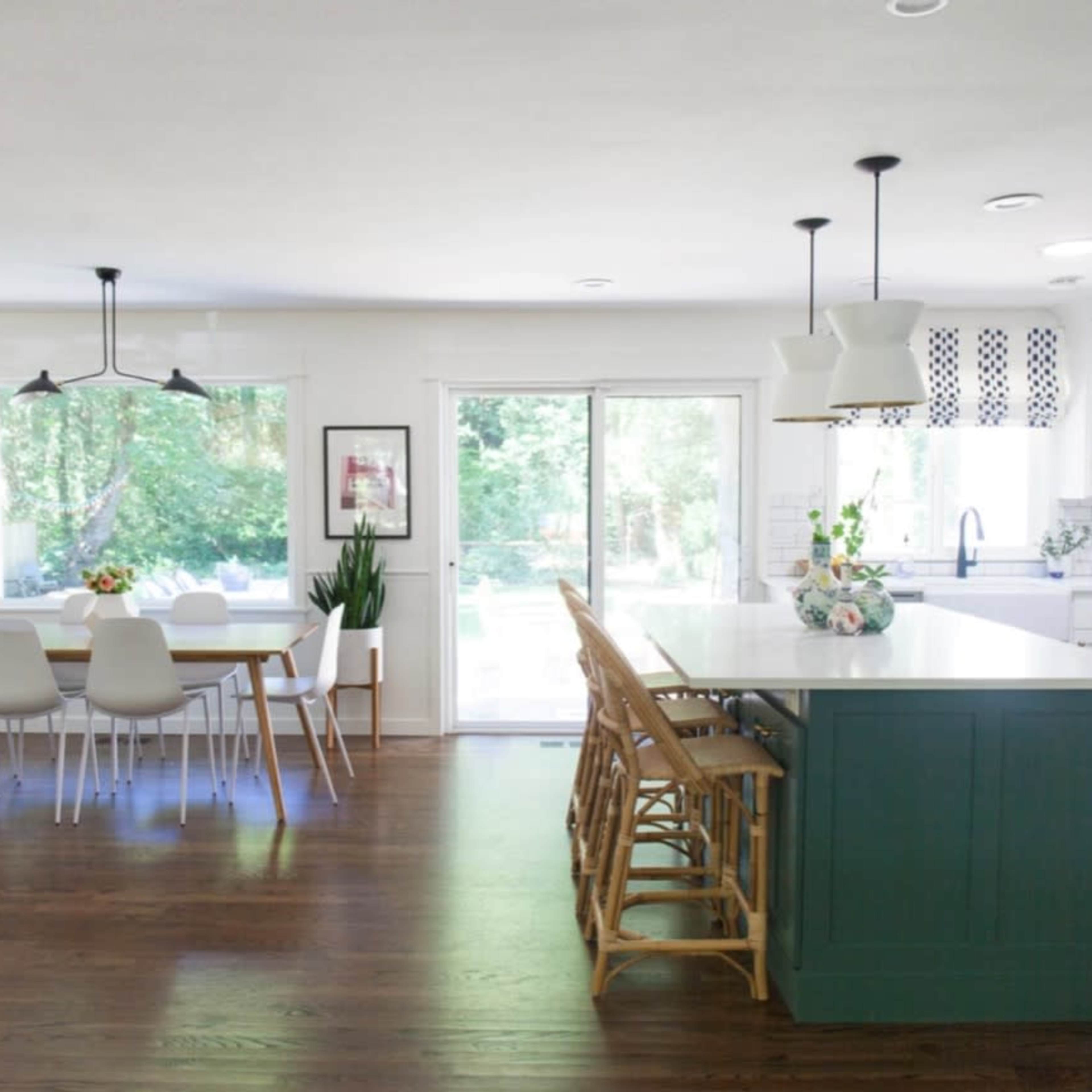 The kitchen features a large island with bar seating, a dining table, and a sliding glass door leading to a backyard, all surrounded by white cabinetry and natural wood floors.