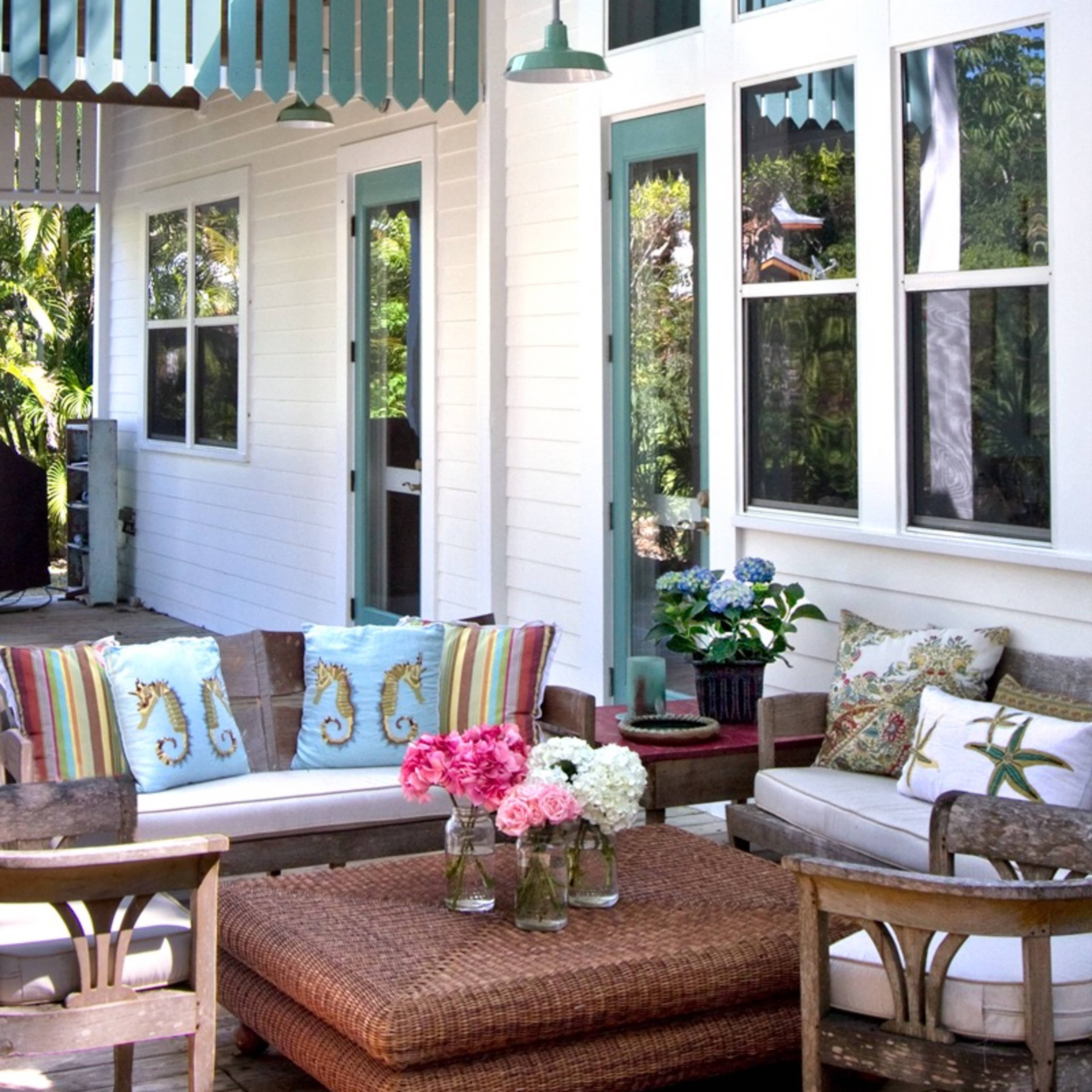 A patio features a seating area with wicker furniture, colorful cushions, and a coffee table surrounded by plants and large windows.