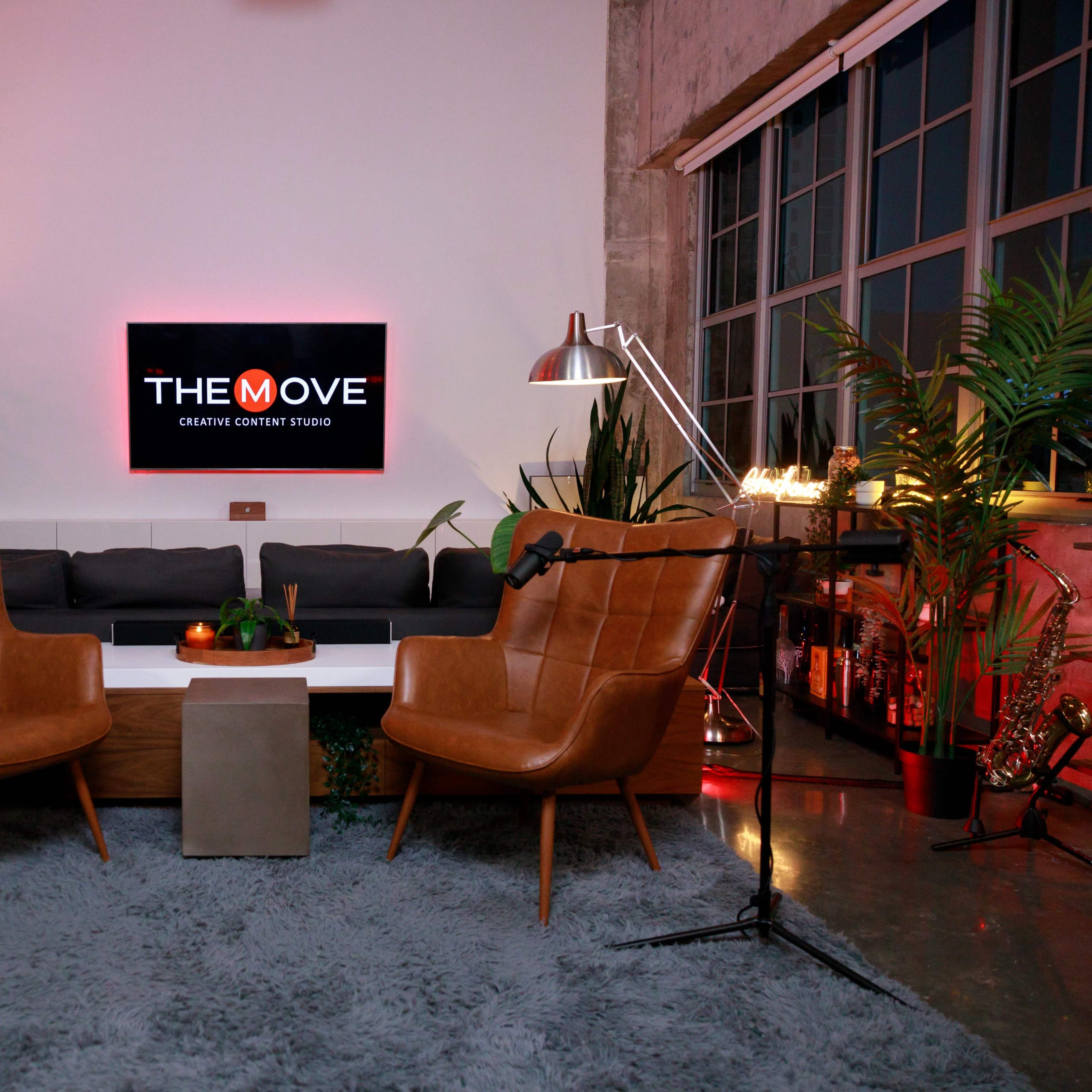 A modern creative content studio featuring two brown leather armchairs facing a white coffee table, with a large screen displaying "THE MOVE" in the background and plants adorning the space.