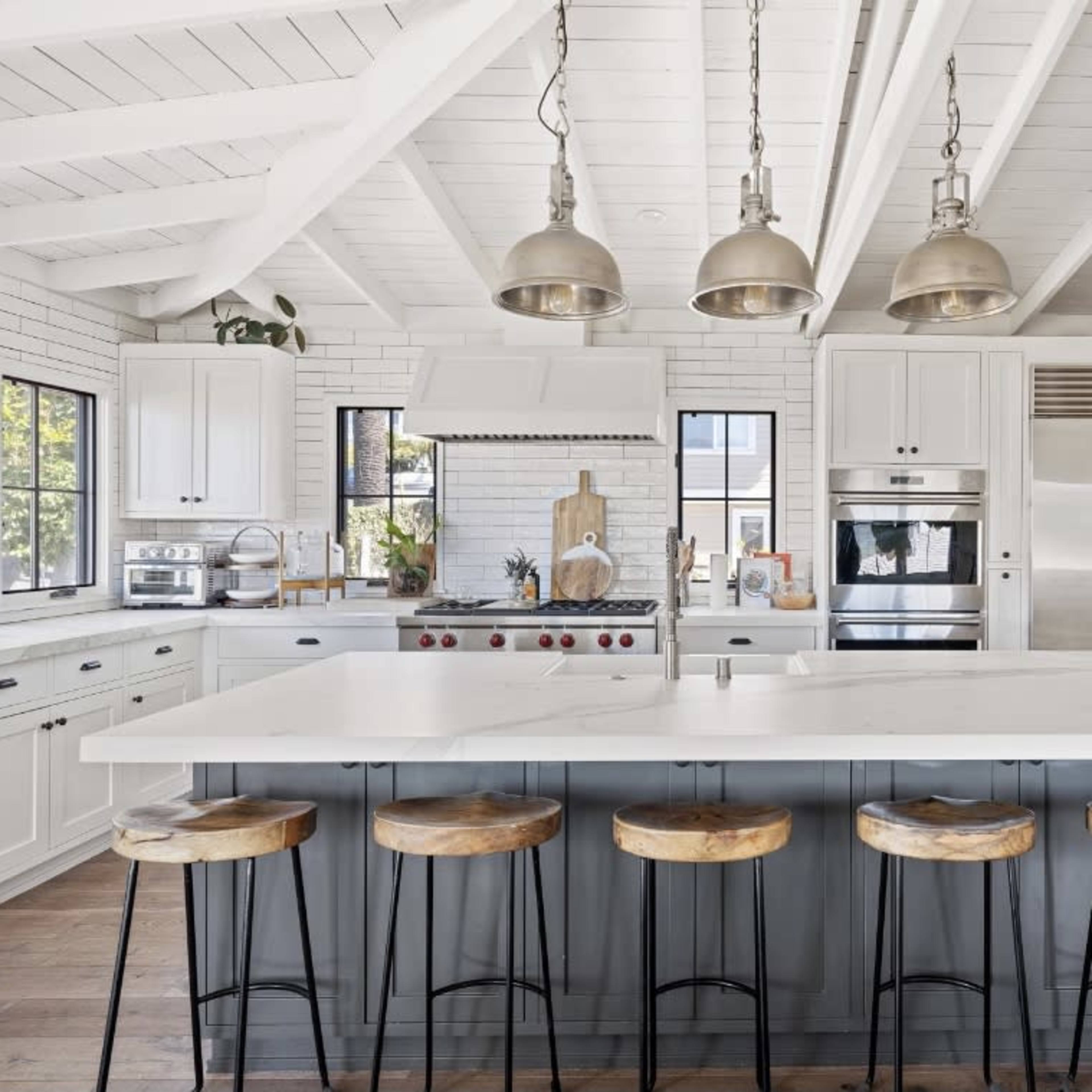 A modern kitchen with a white ceiling, wooden beams, a large island with bar stools, and stainless steel appliances.
