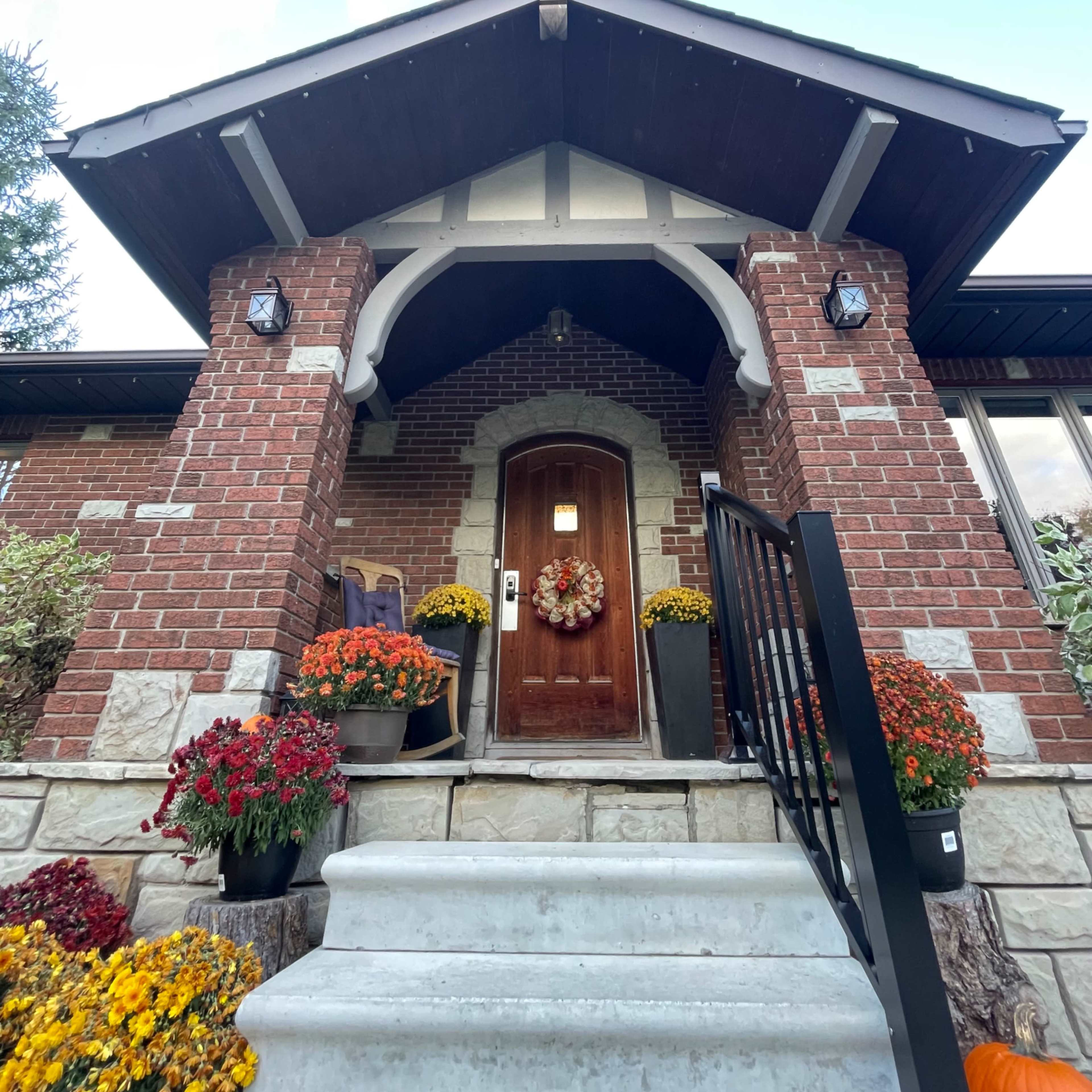 The entrance of a brick house featuring a large wooden door under a decorative archway, flanked by colorful flowerpots and decorative lighting.
