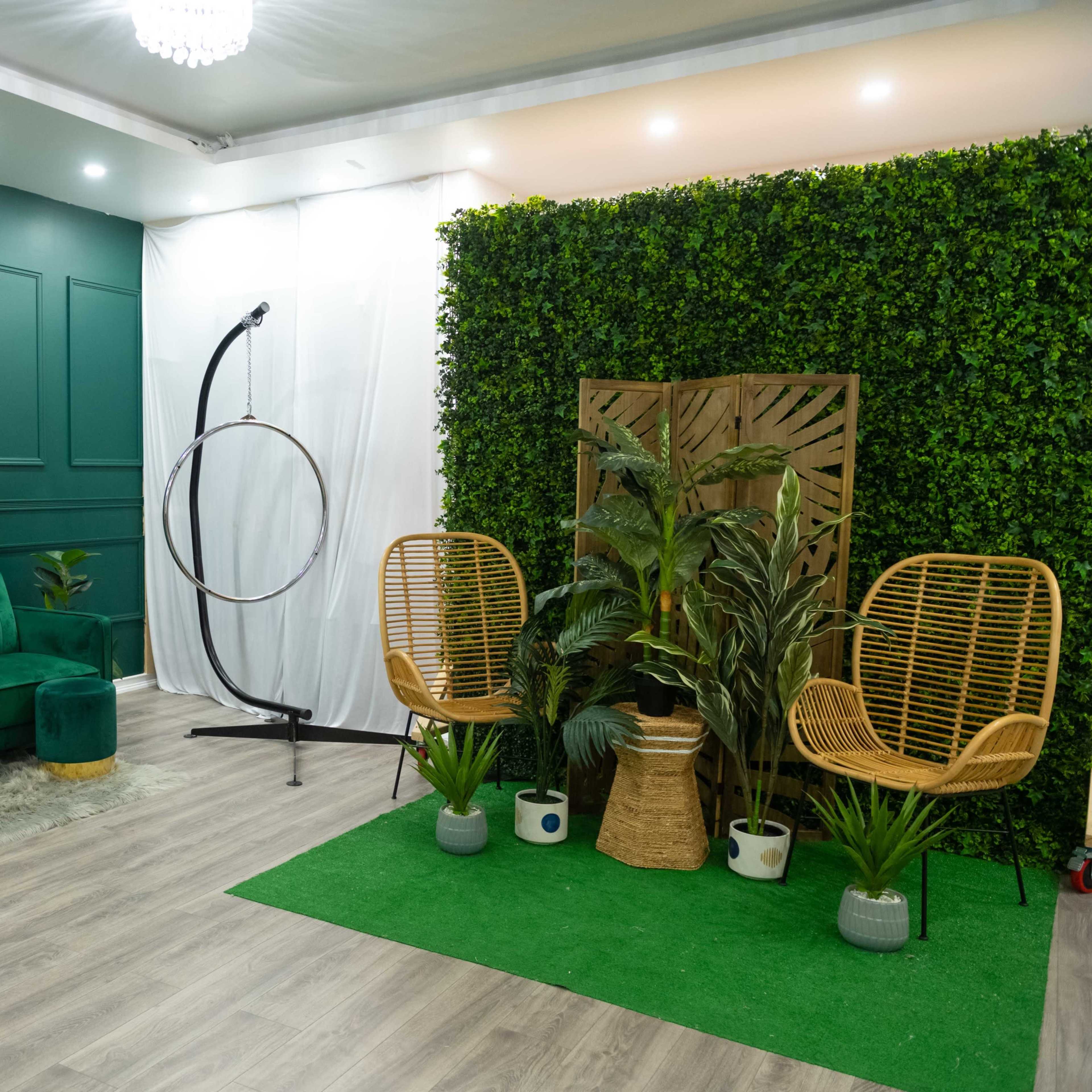 A modern indoor space featuring a green tufted sofa, two rattan chairs, a hoop structure, and decorative plants against a backdrop of artificial greenery.