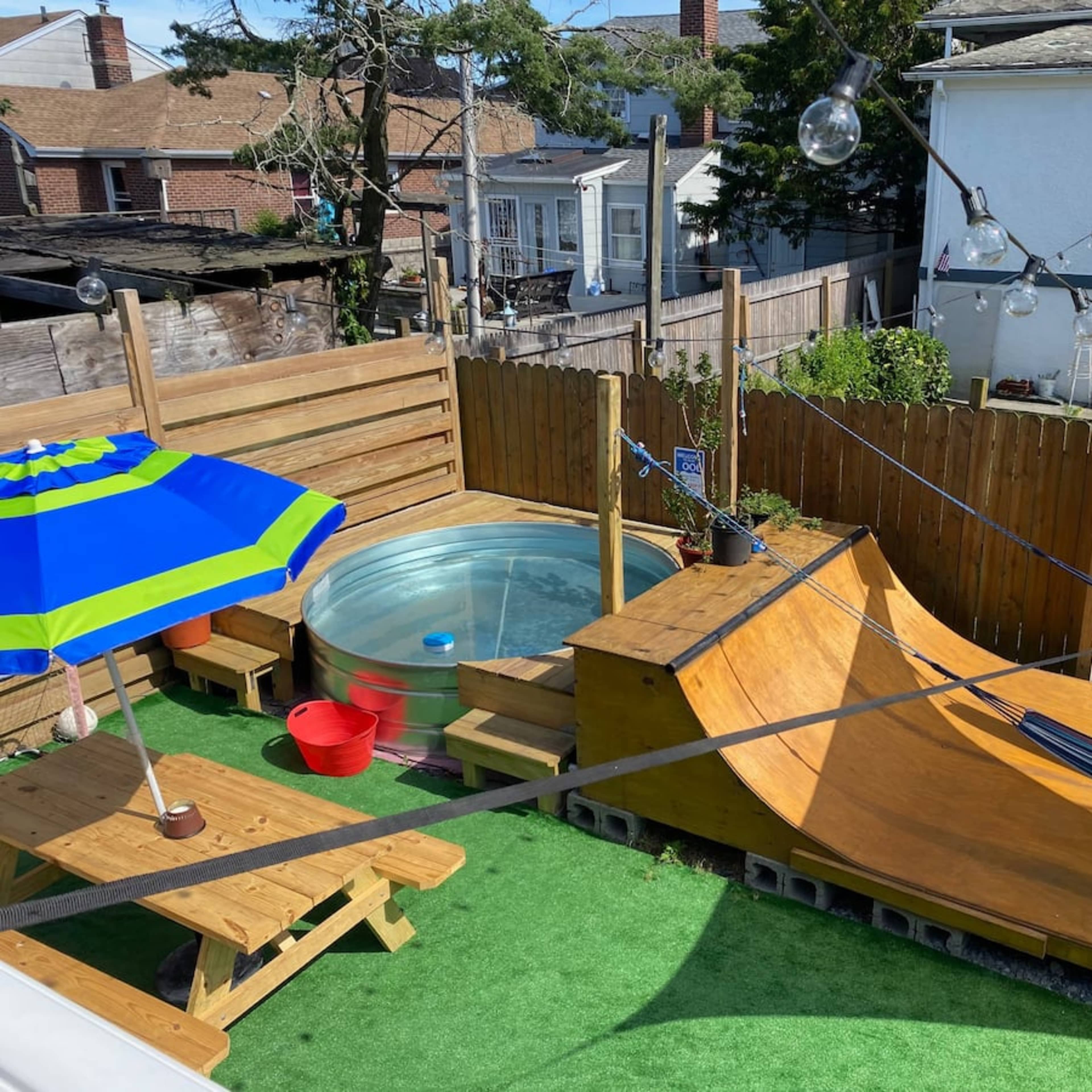 A backyard featuring a small wooden ramp, a hot tub, a picnic table, and a shaded seating area, all surrounded by green artificial grass and wooden fencing.