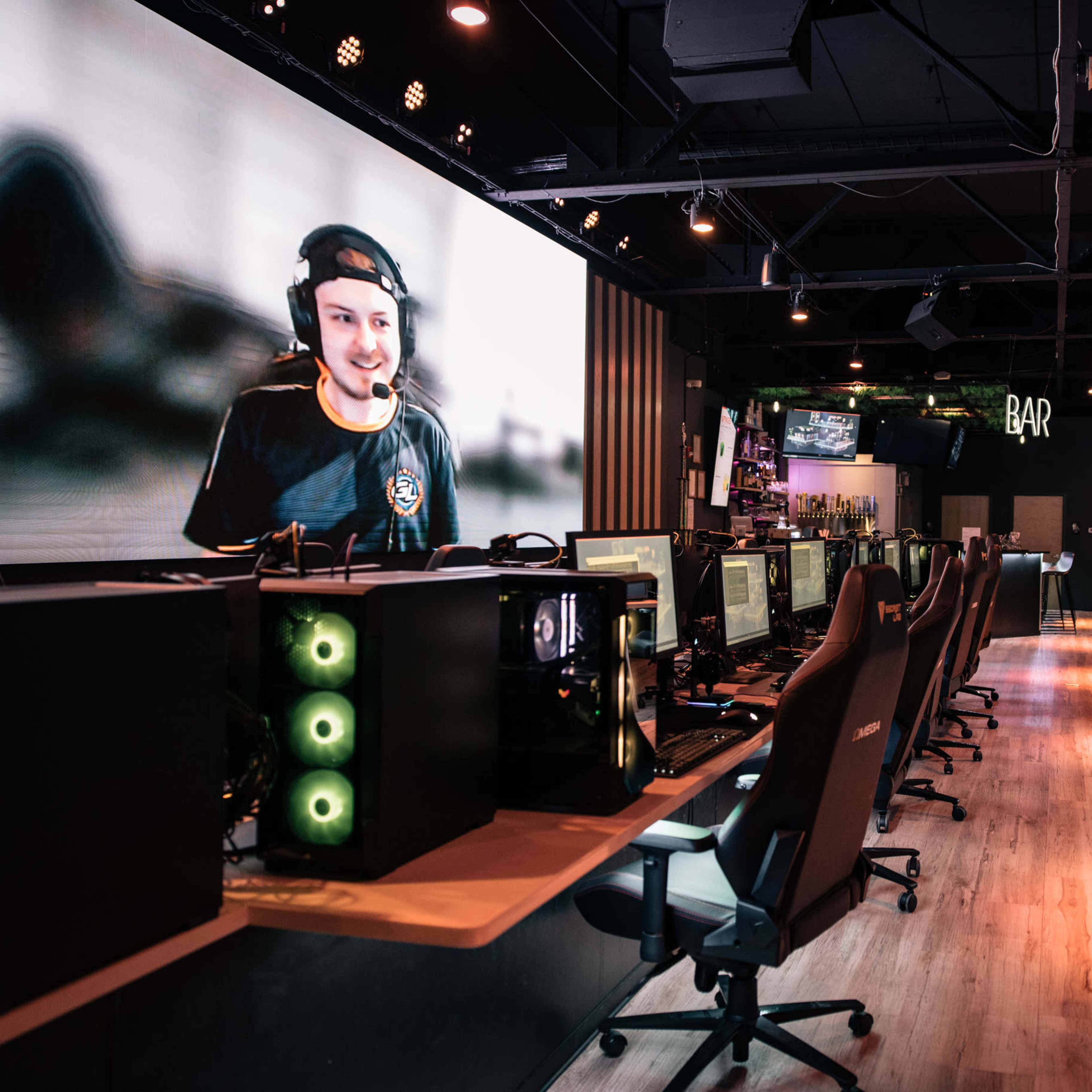 A gaming lounge with rows of computer gaming stations, a large screen displaying a player's face, and various gaming setups.