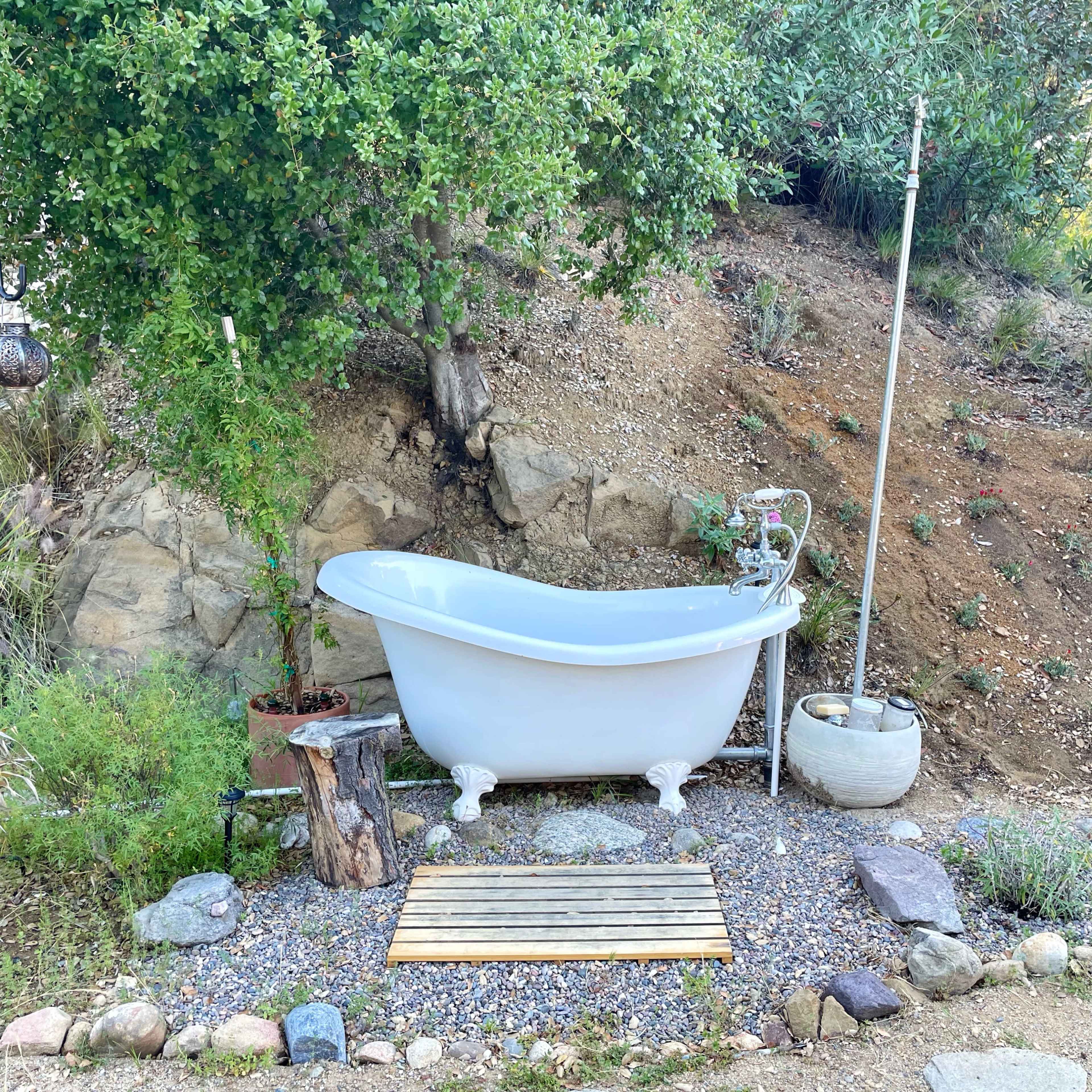 A freestanding white bathtub sits on a wooden platform surrounded by rocks and greenery in an outdoor setting.