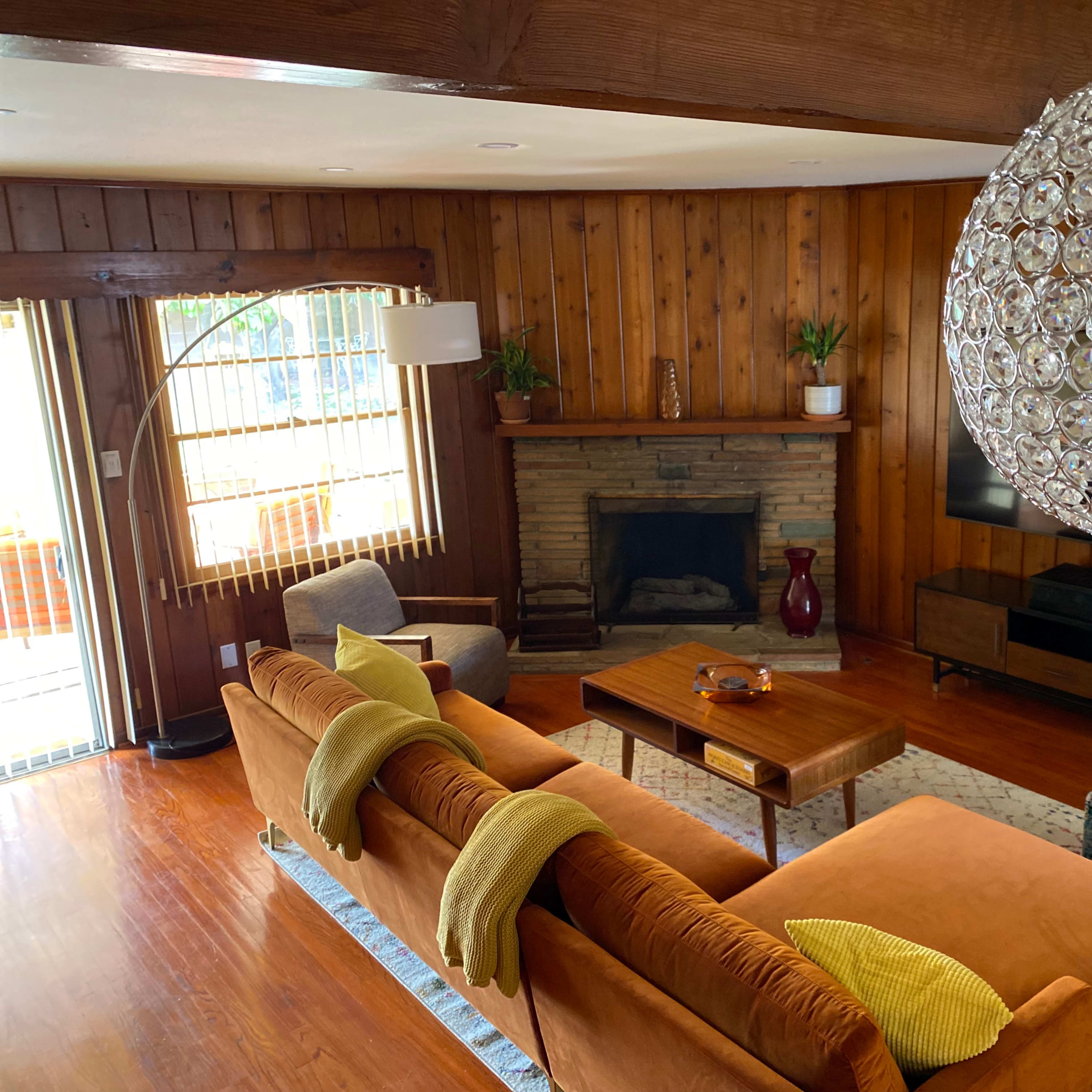 A spacious living room with wood paneling, a stone fireplace, a large round chandelier, and a sofa adorned with yellow throw blankets.