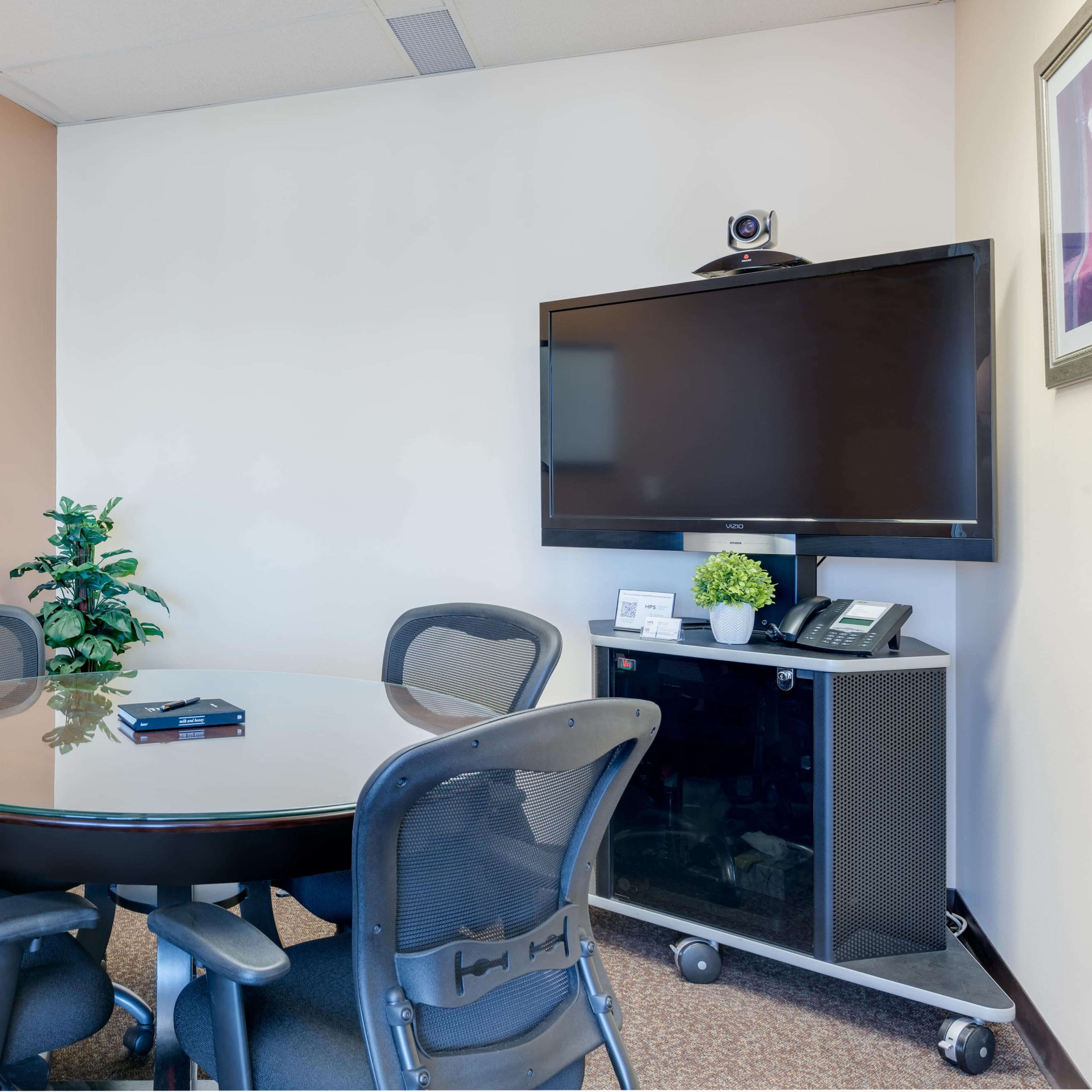 A small conference room with a round glass table, four black chairs, a potted plant, a wall-mounted television, and a whiteboard.