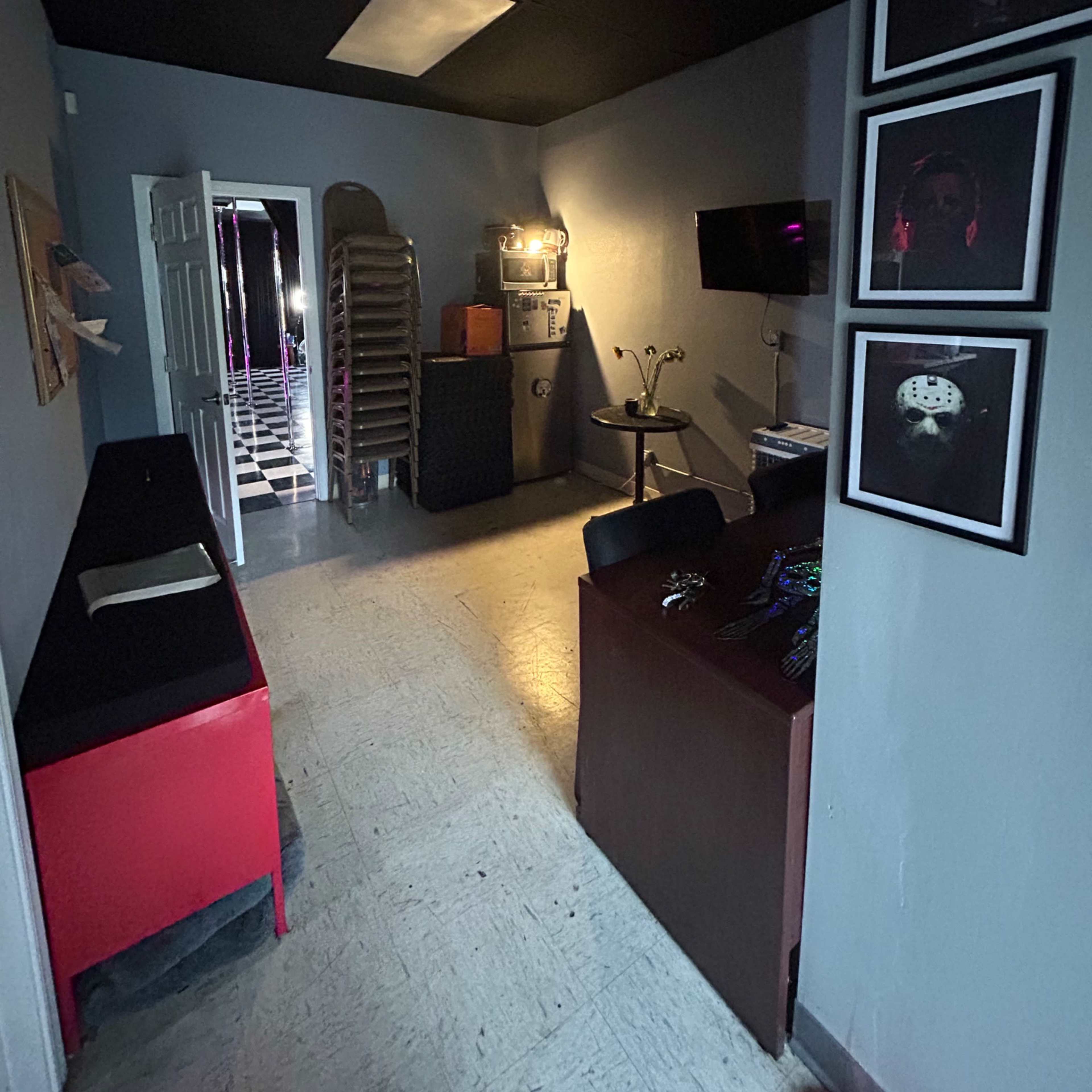 A narrow, dimly lit hallway leading to a room with a checkered floor, featuring various furniture pieces and a door at the end.