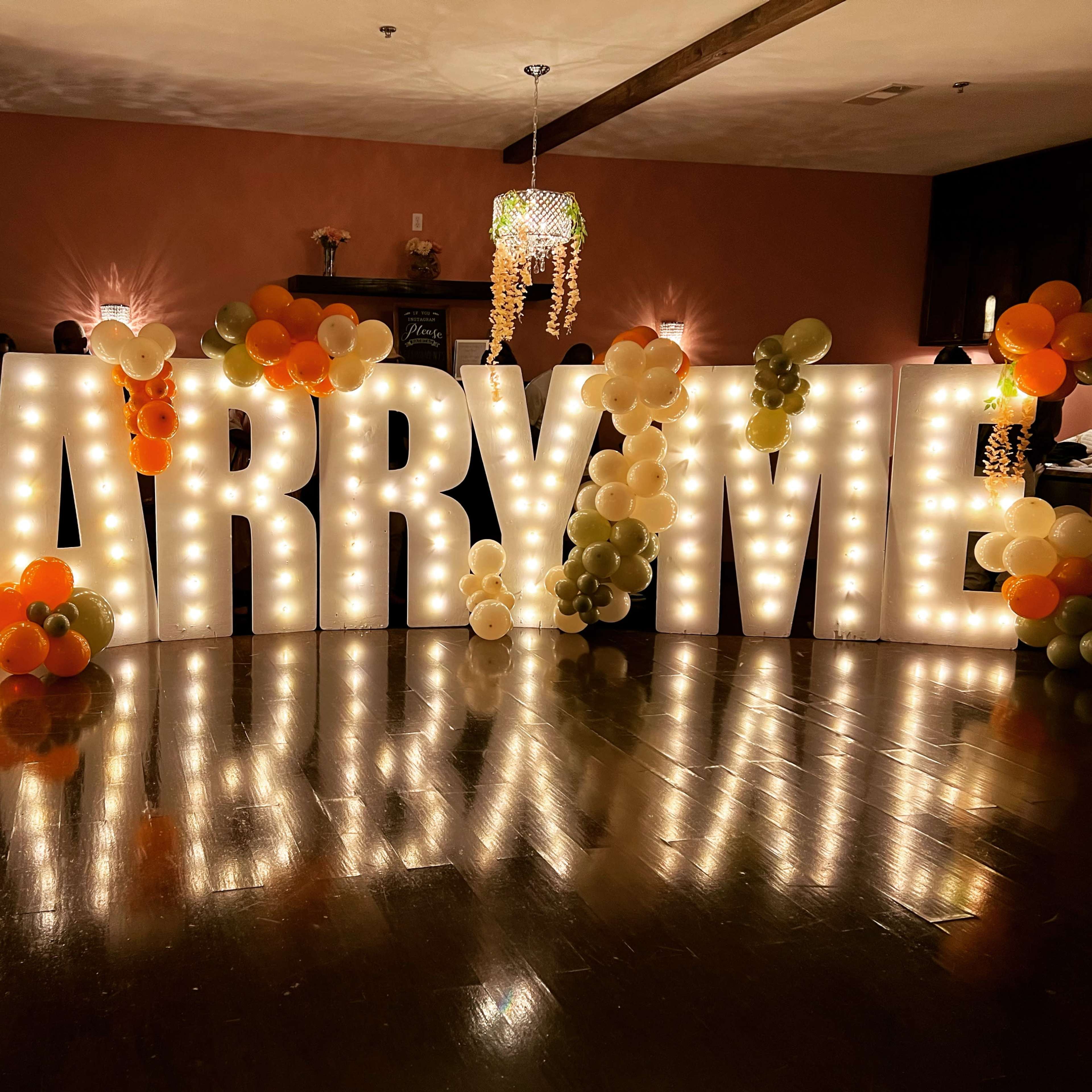 The scene features large, illuminated letters spelling "MARRY ME," decorated with an array of balloons in various colors.