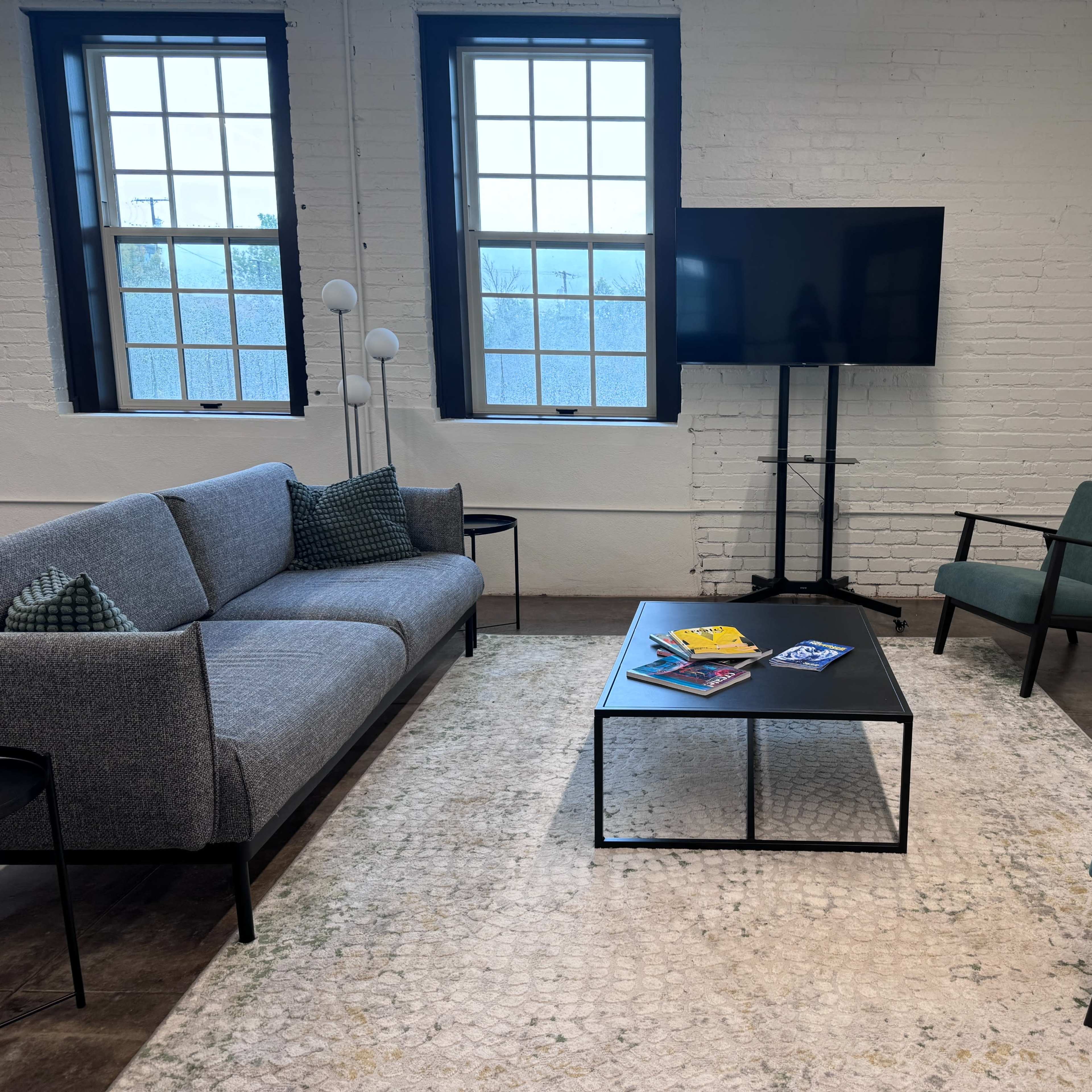 A modern lounge area with a gray sofa, two green chairs, a black coffee table, a television on a stand, and large windows allowing natural light.