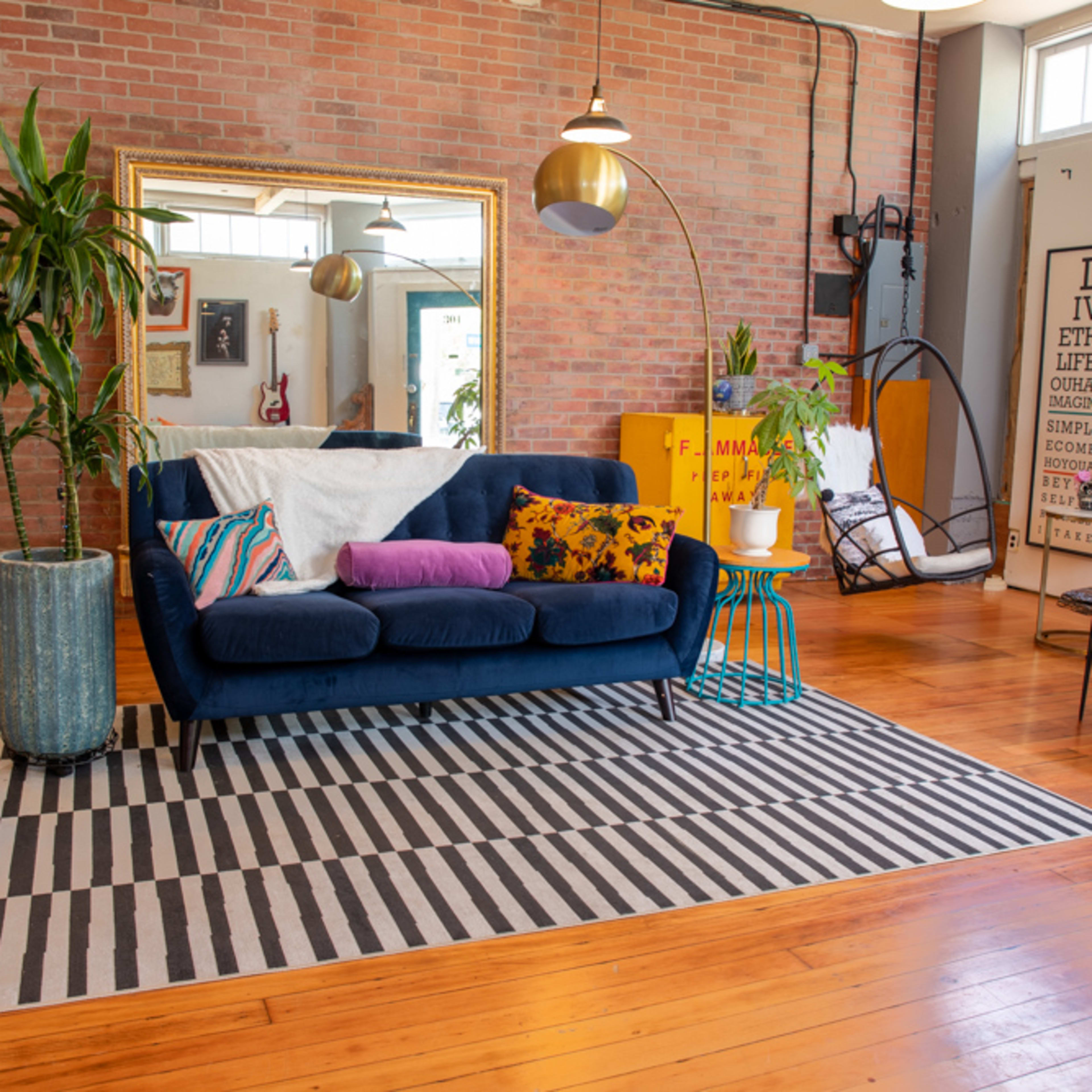 A bright living room features a blue sofa with colorful cushions, a large mirror, a potted plant, and a striped rug on polished wooden flooring.
