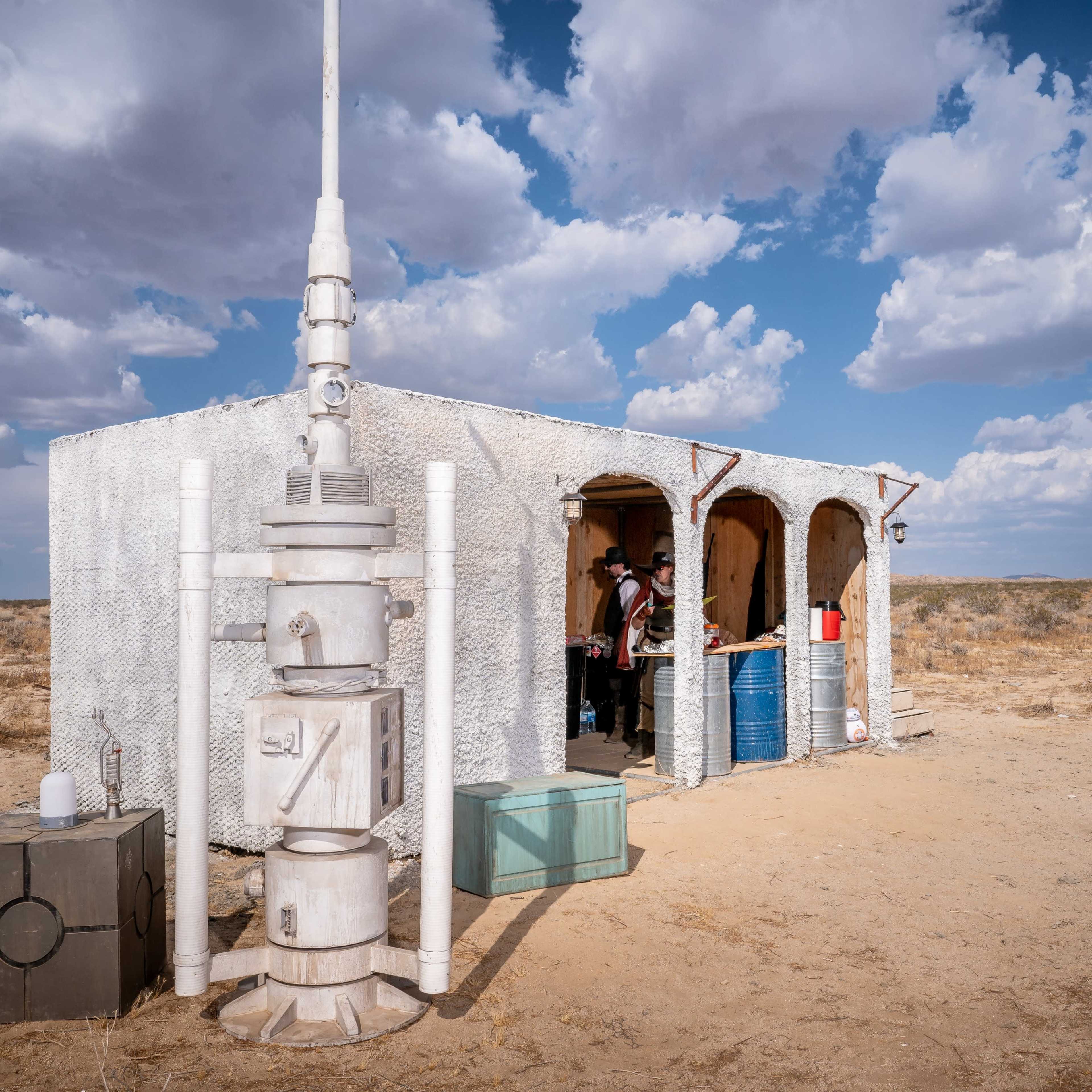 A small, white, adobe-style building with three arched doorways sits in a barren landscape under a cloudy sky.
