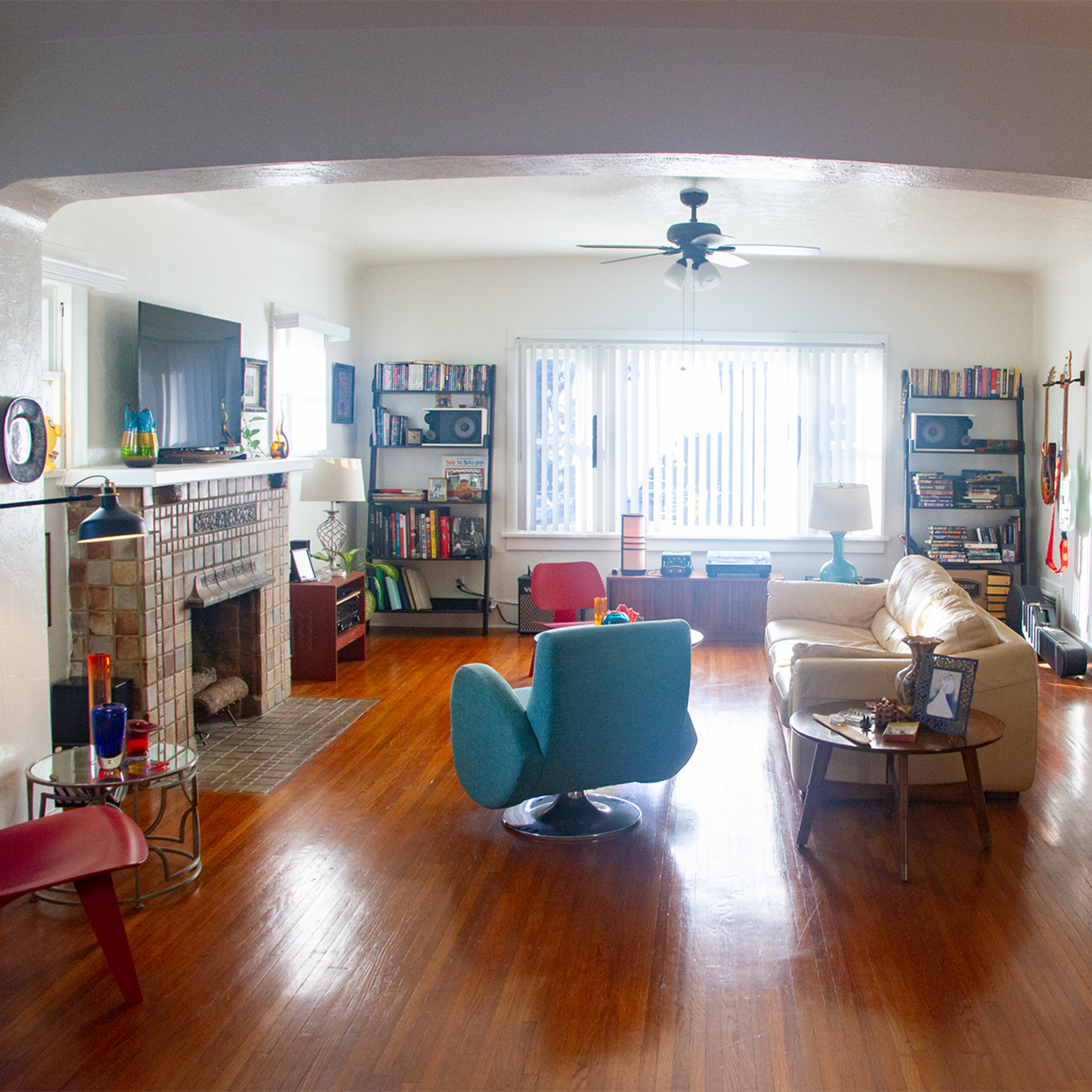 The living room features a fireplace, bookshelves, and a mix of seating arrangements, including a blue chair and a beige sofa, with hardwood floors and a view of windows in the background.