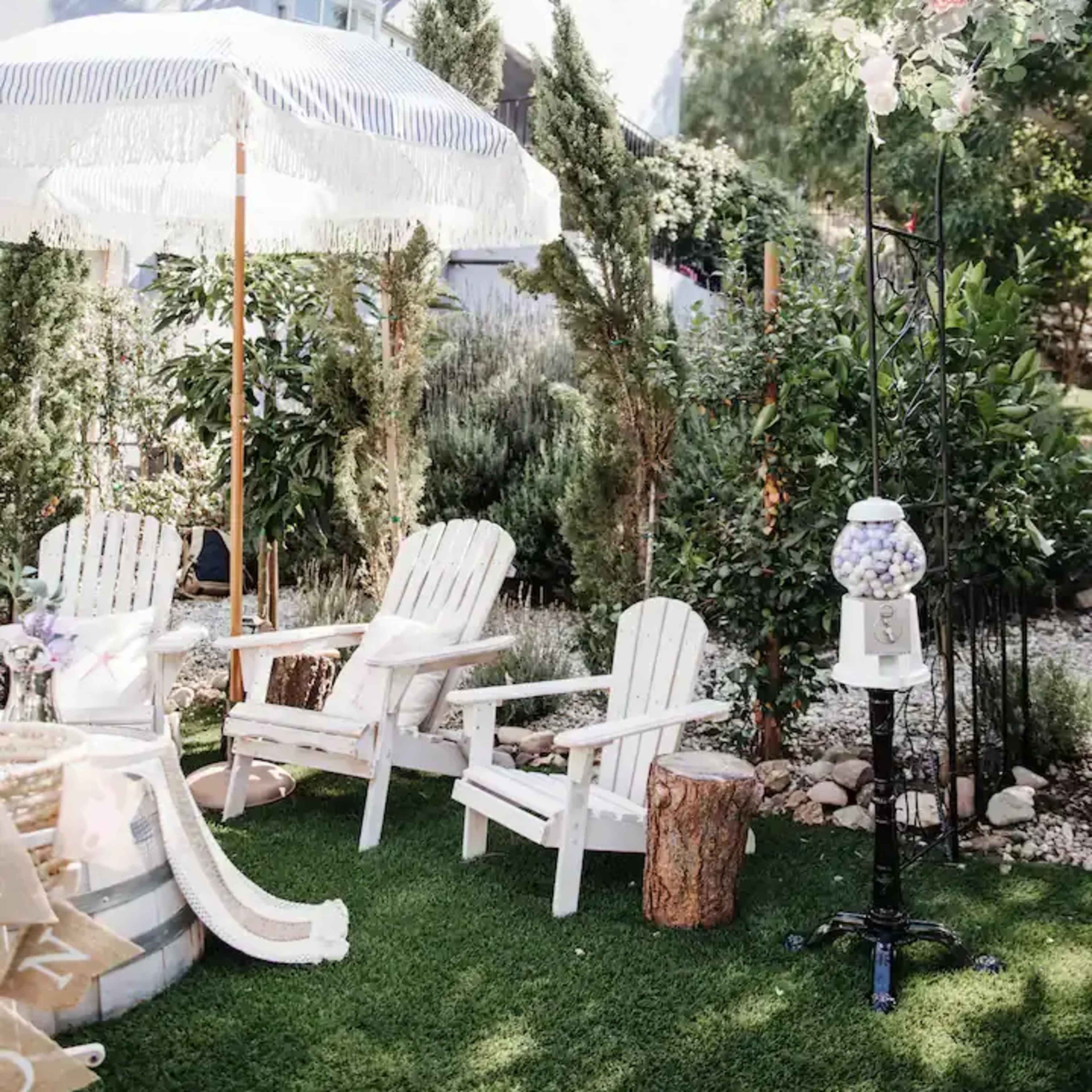 A garden area with white Adirondack chairs, a decorative umbrella, and a gumball machine on a grassy surface surrounded by lush greenery.