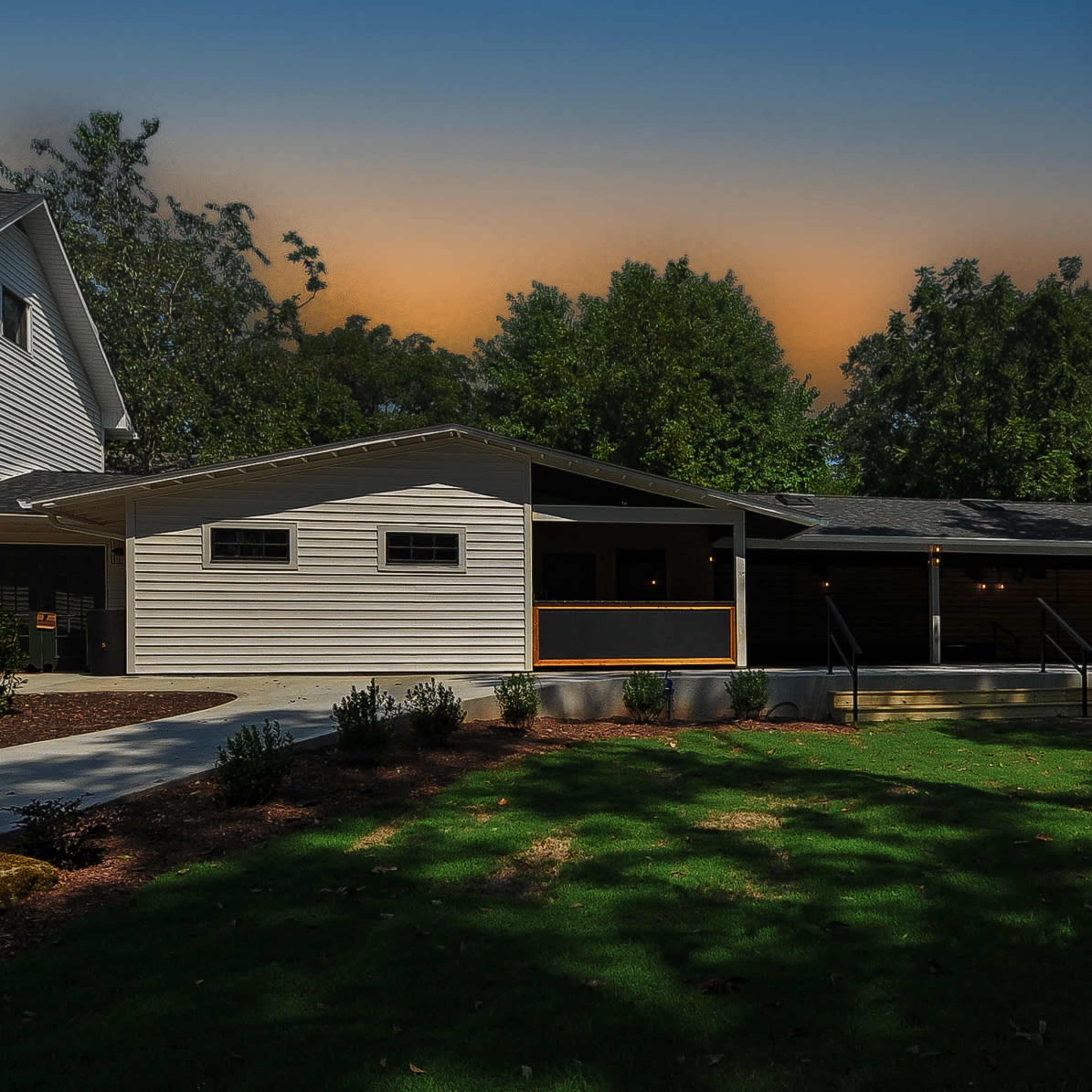 A modern single-story house with a landscaped yard and a wooden deck surrounded by trees at sunset.