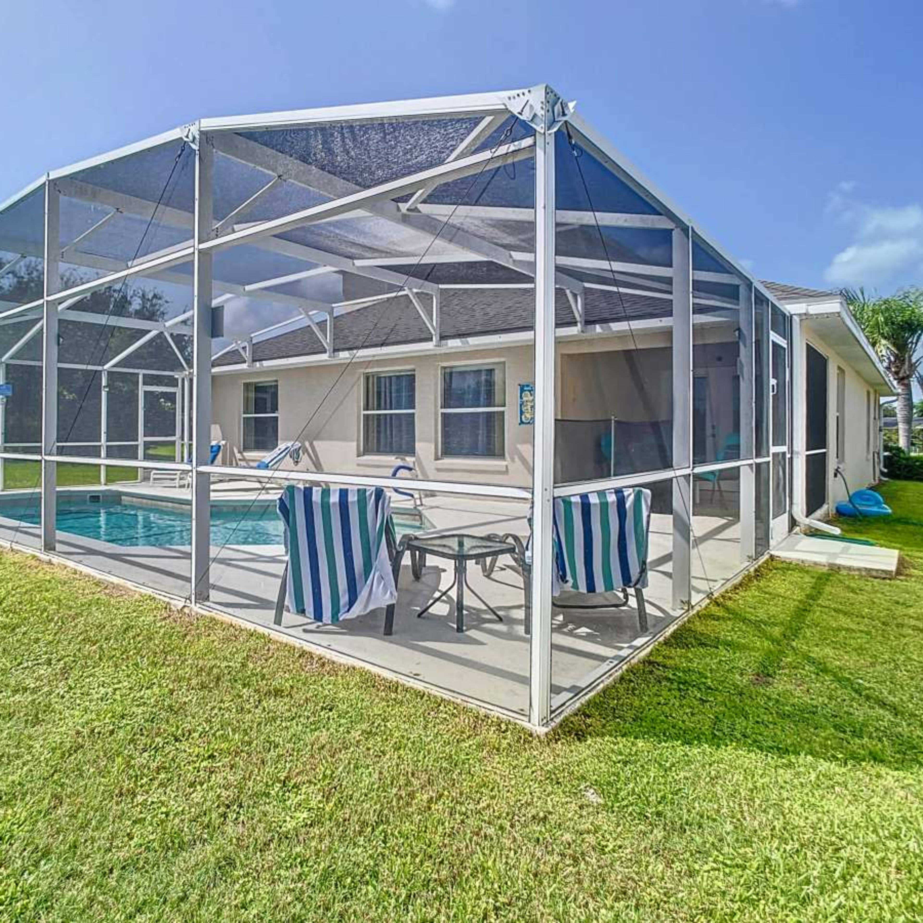 A screened-in pool area with lounge chairs and tables is situated beside a house, surrounded by green grass and palm trees.