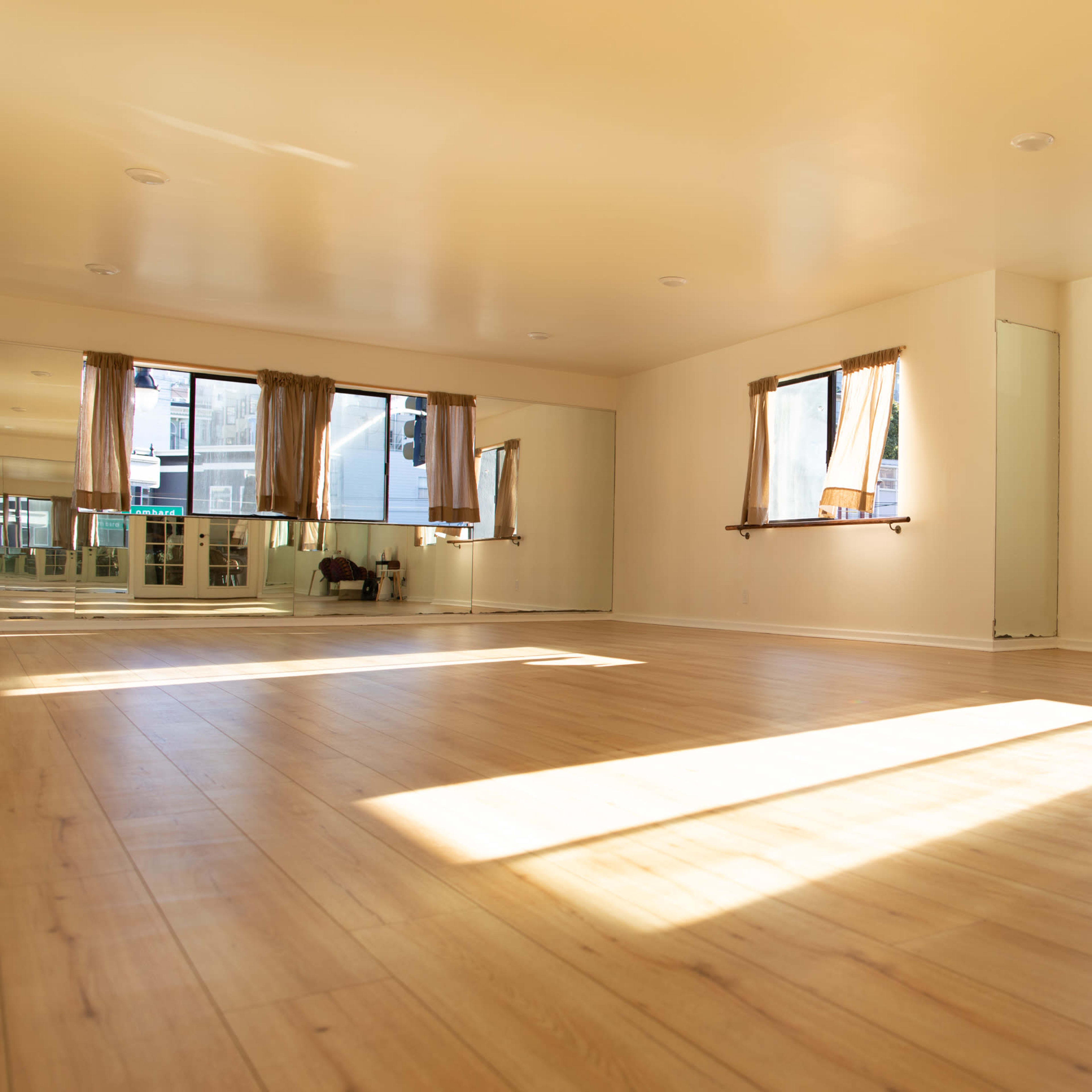 A spacious room with wooden floors, two large windows dressed with sheer curtains, and ample natural light streaming in.