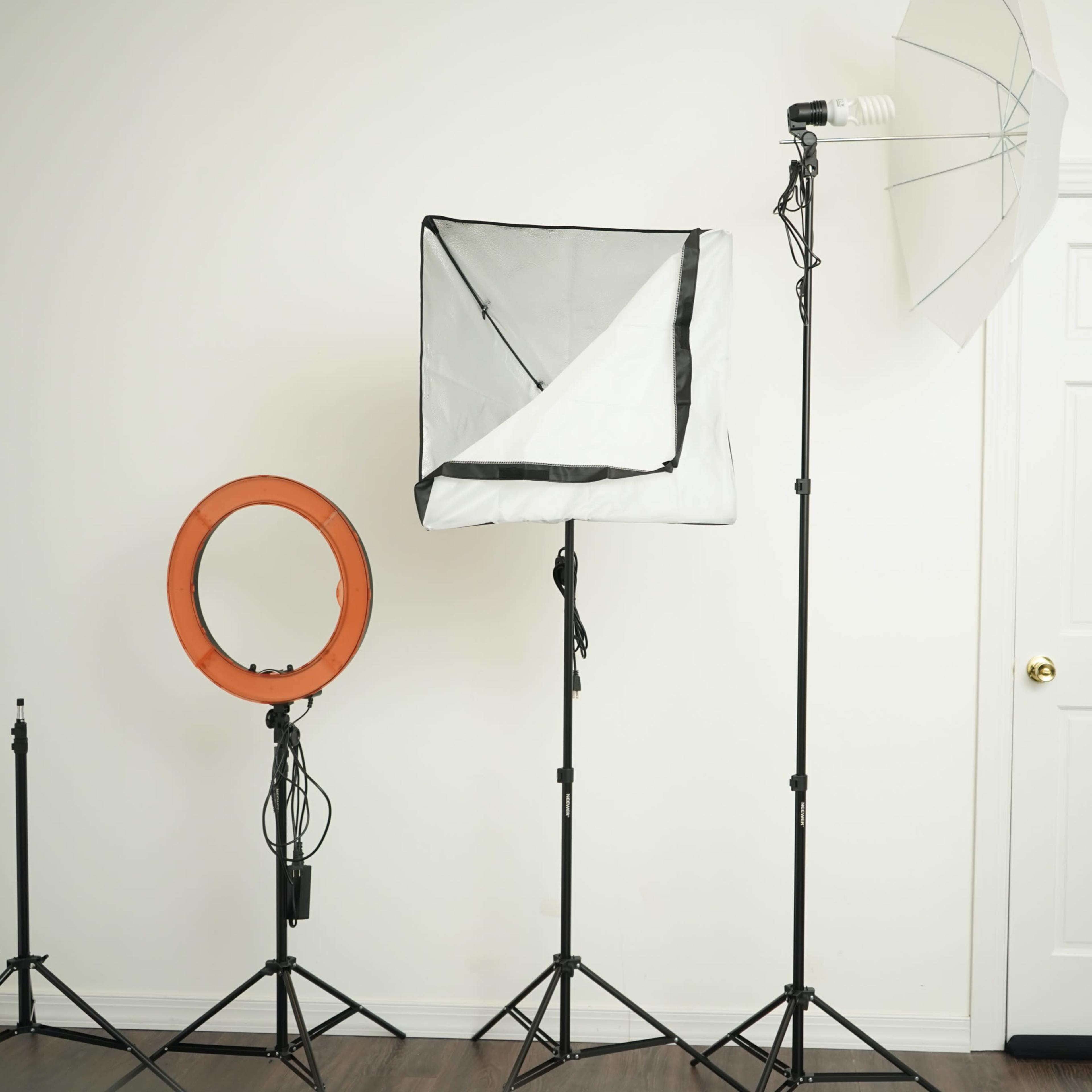 Photography lighting equipment, including a ring light, a softbox, and an umbrella, arranged in front of a white wall and a closed door.