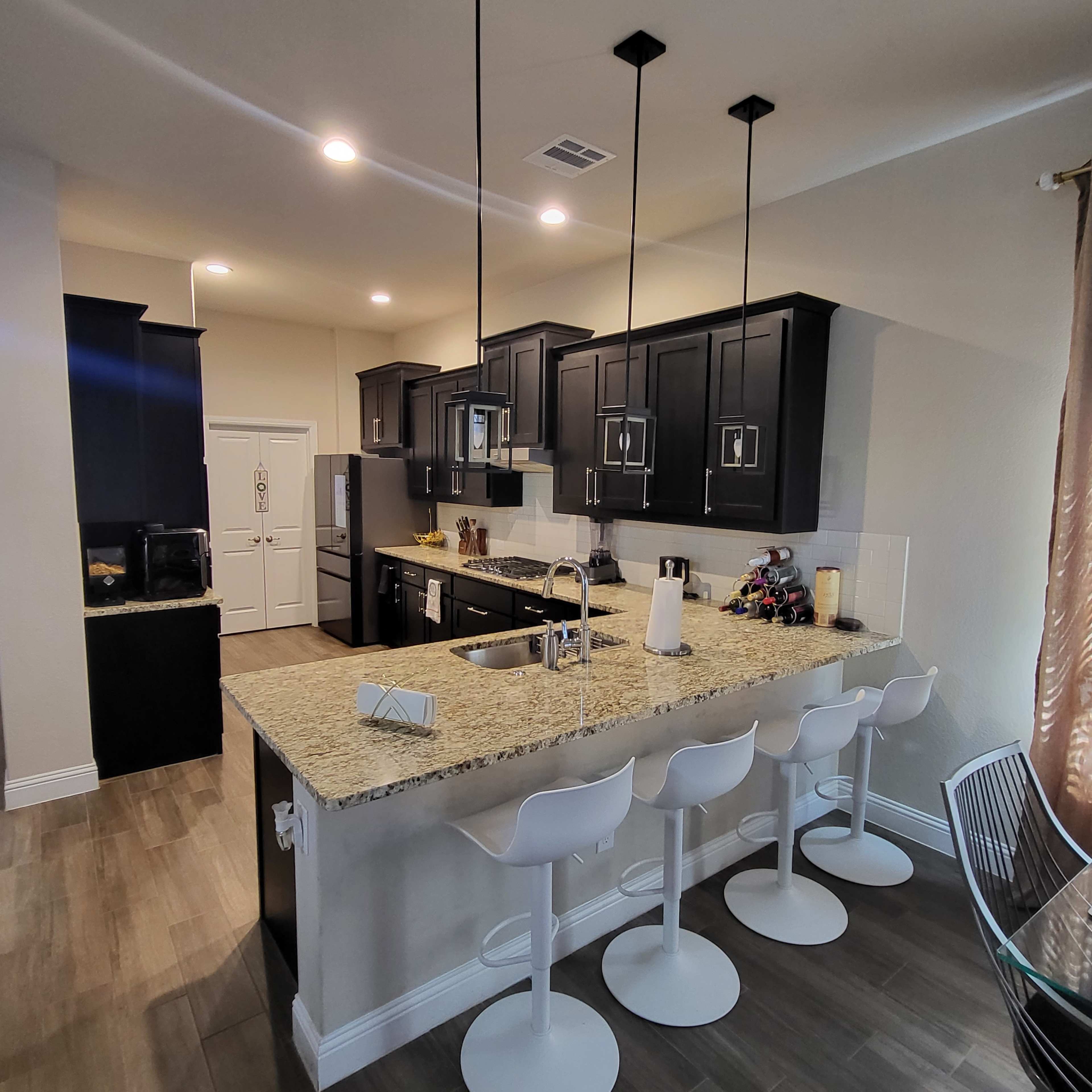 A modern kitchen with dark cabinets, a central island with four white bar stools, and stainless steel appliances.