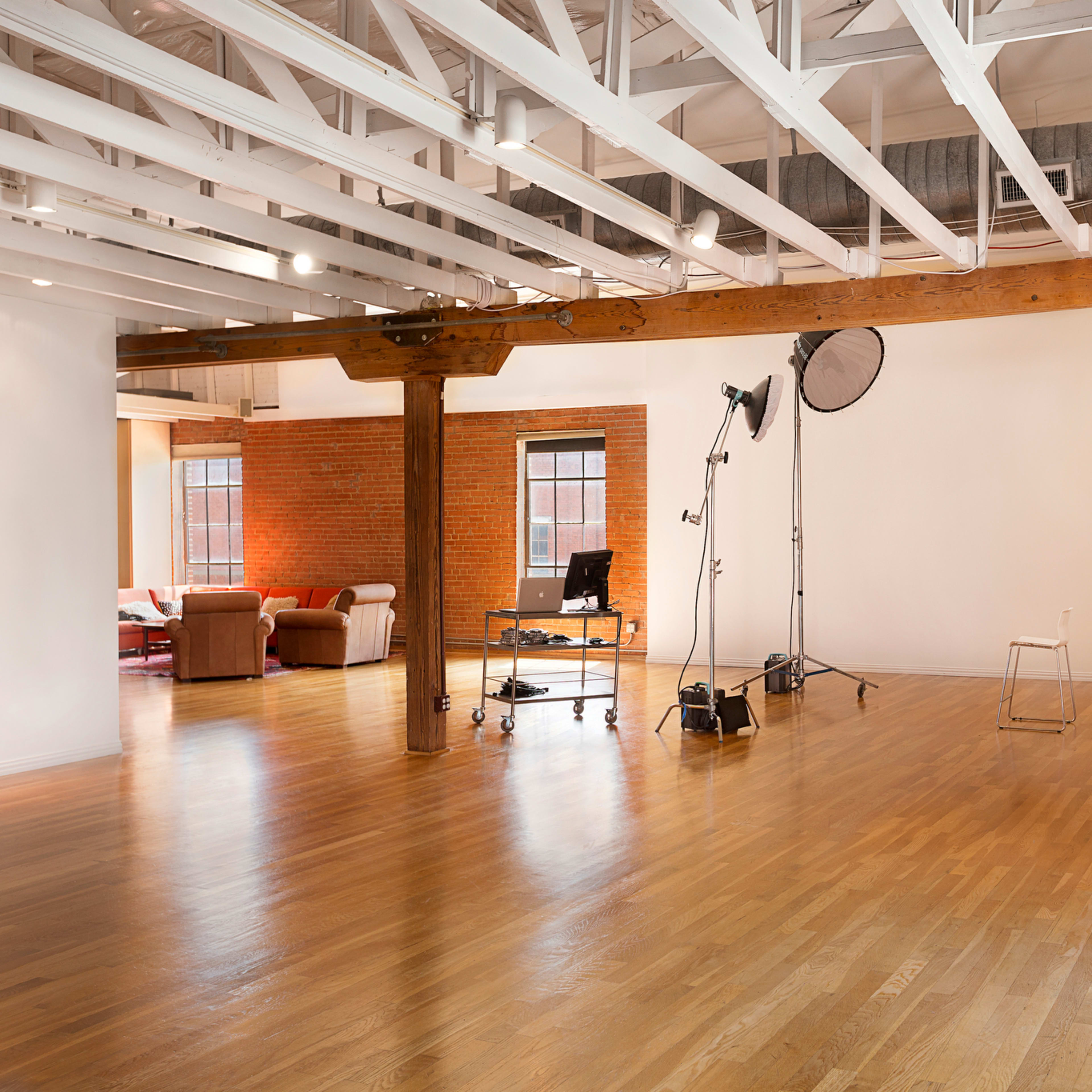 An open space with hardwood floors, exposed wooden beams, and studio lighting equipment set up on one side, while a cozy seating area is visible in the background.