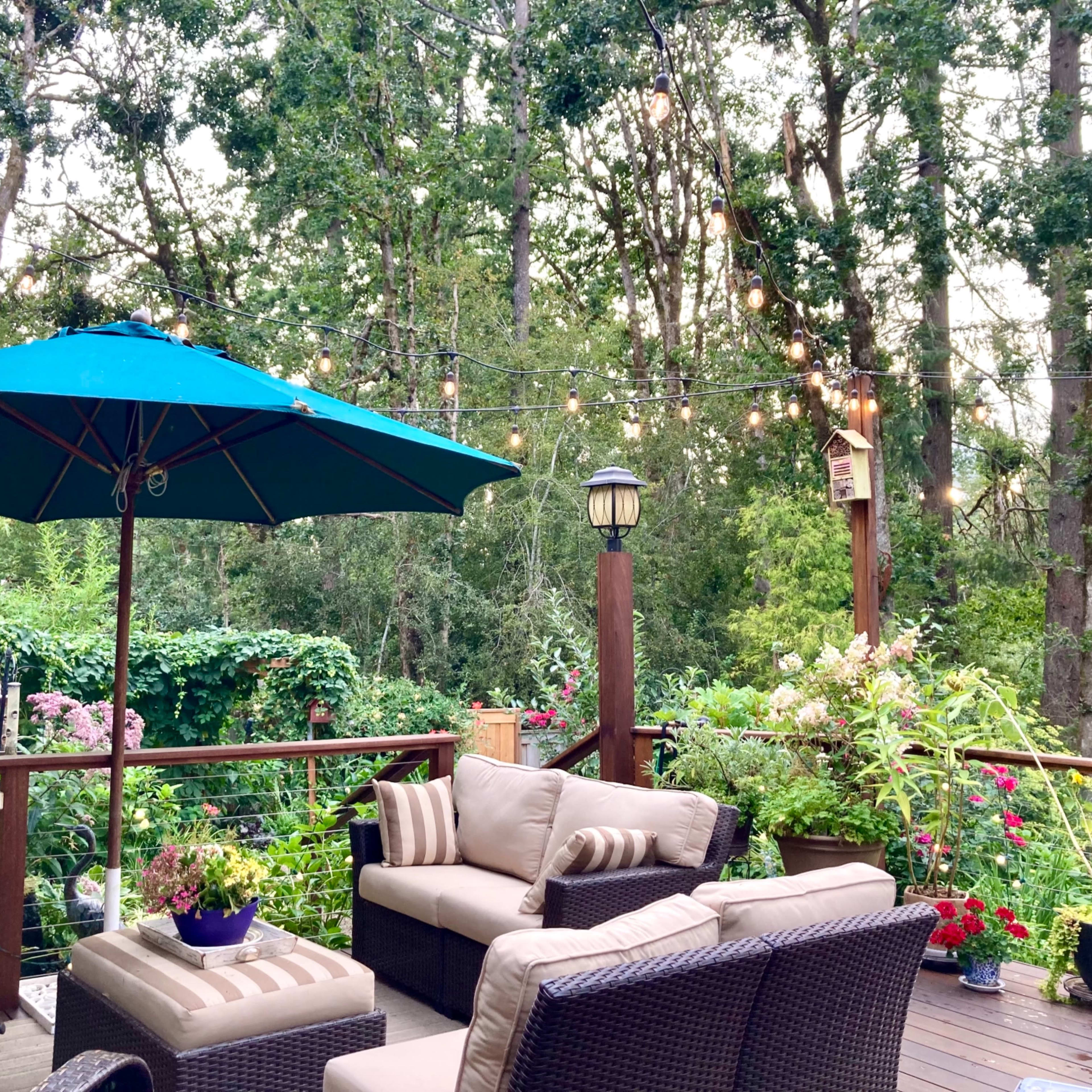 A patio area is furnished with a seating arrangement under a large umbrella, surrounded by greenery and string lights.