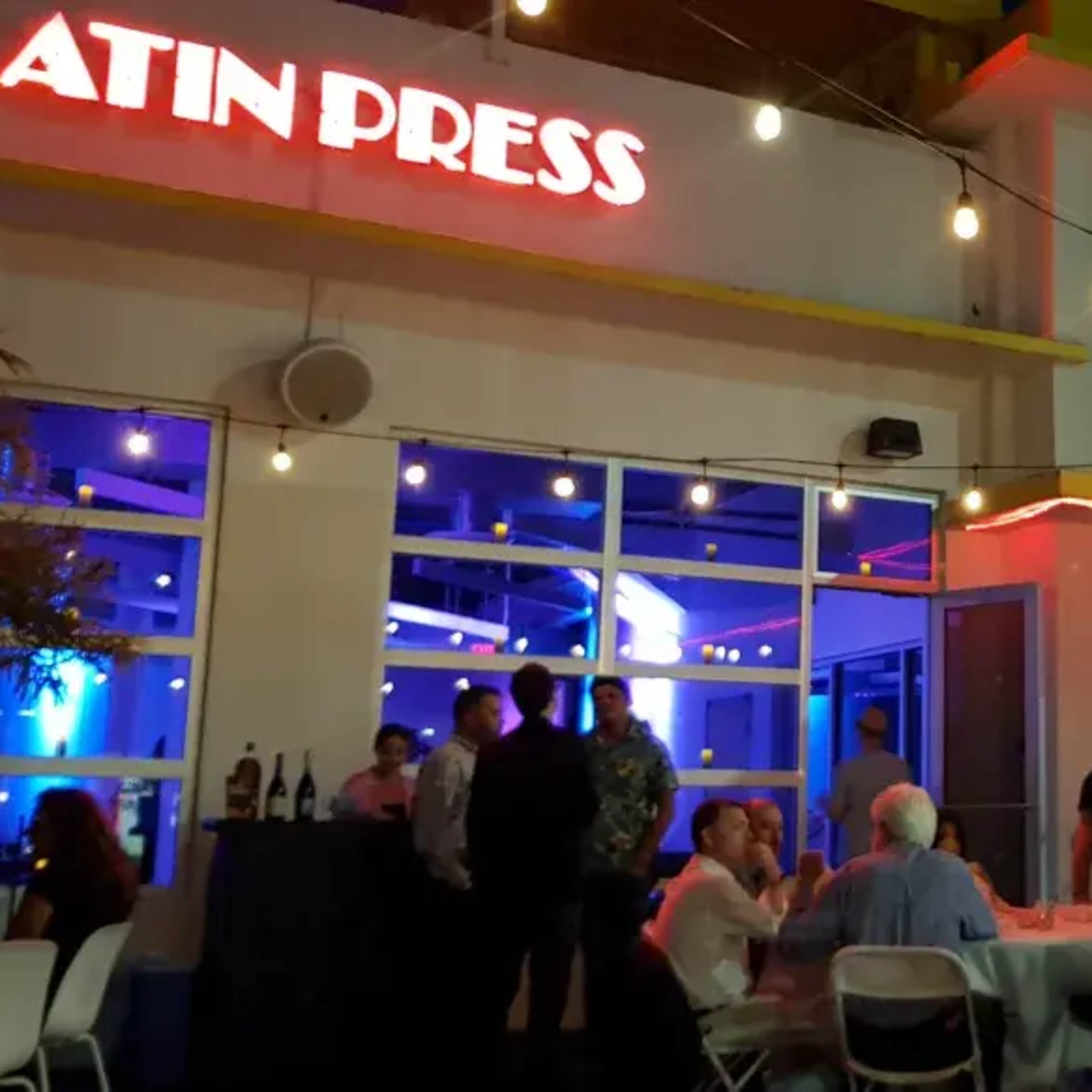A gathering takes place outside a building labeled "Latin Press," with attendees seated at tables and mingling under string lights.