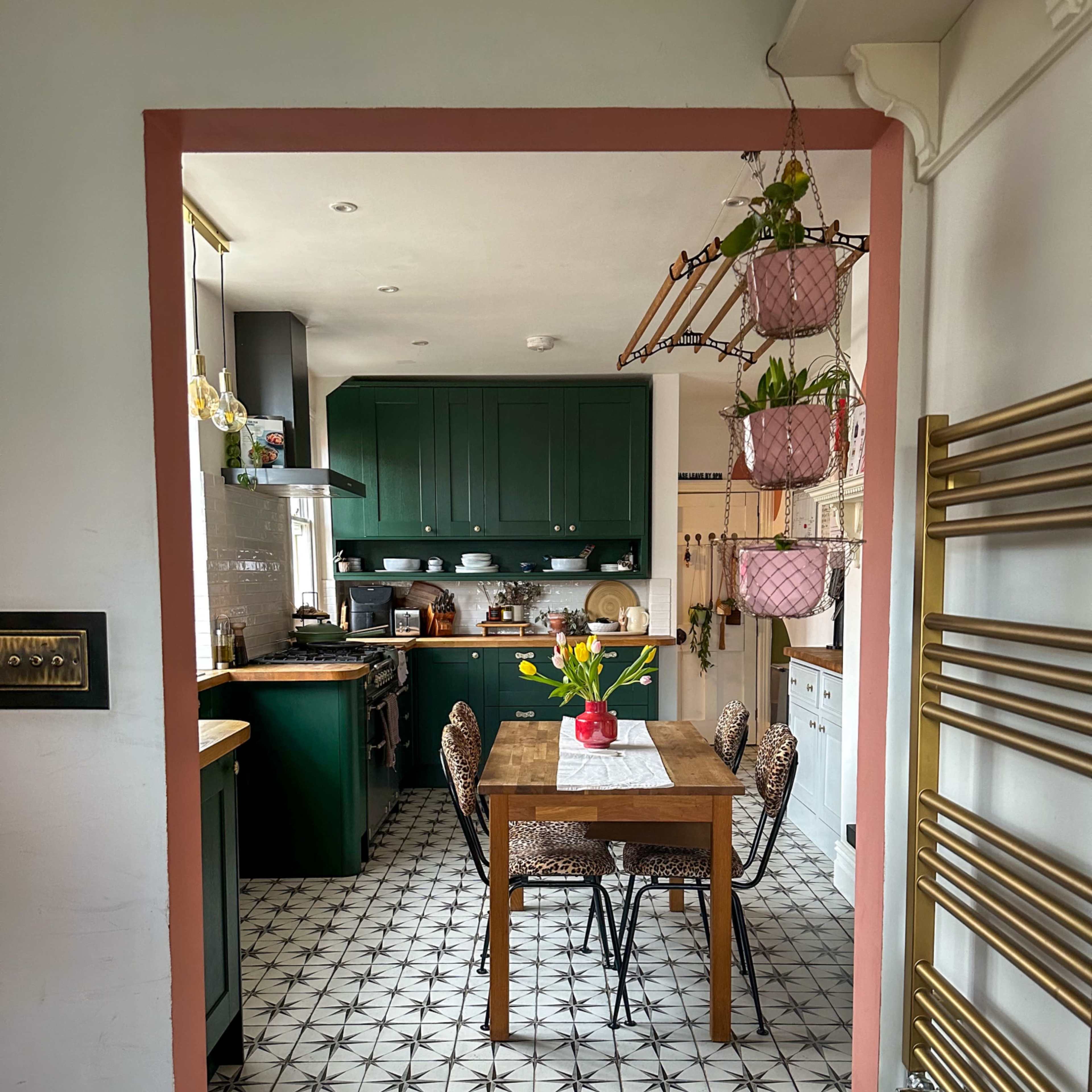 A kitchen with dark green cabinetry, a wooden dining table at the center, and patterned tile flooring is visible through an archway.