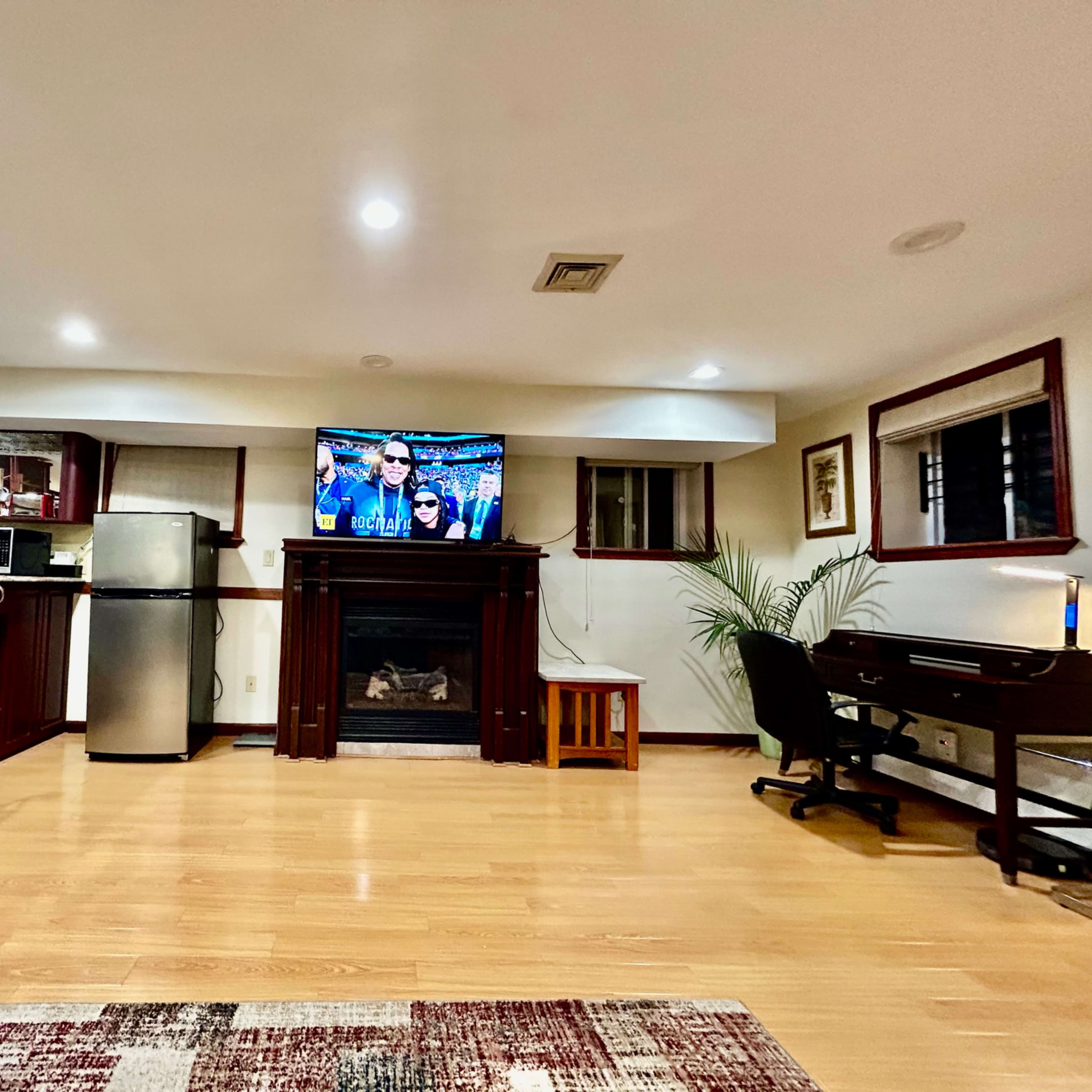 A basement room features a television above a fireplace, a bar area with stools, a desk, and a kitchenette.