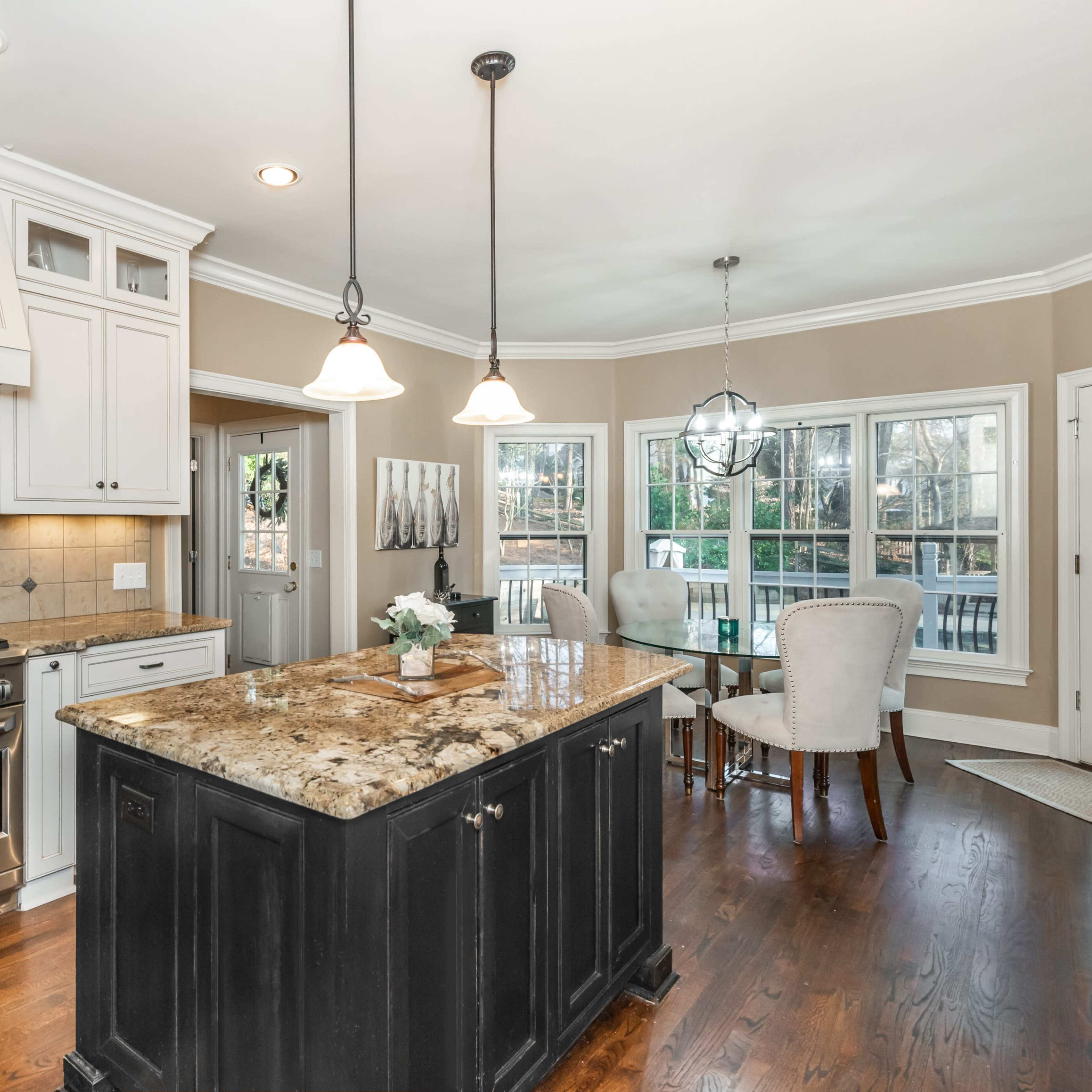 A spacious kitchen features a central island with a dark finish, granite countertops, and stainless steel appliances, surrounded by large windows and a dining area.
