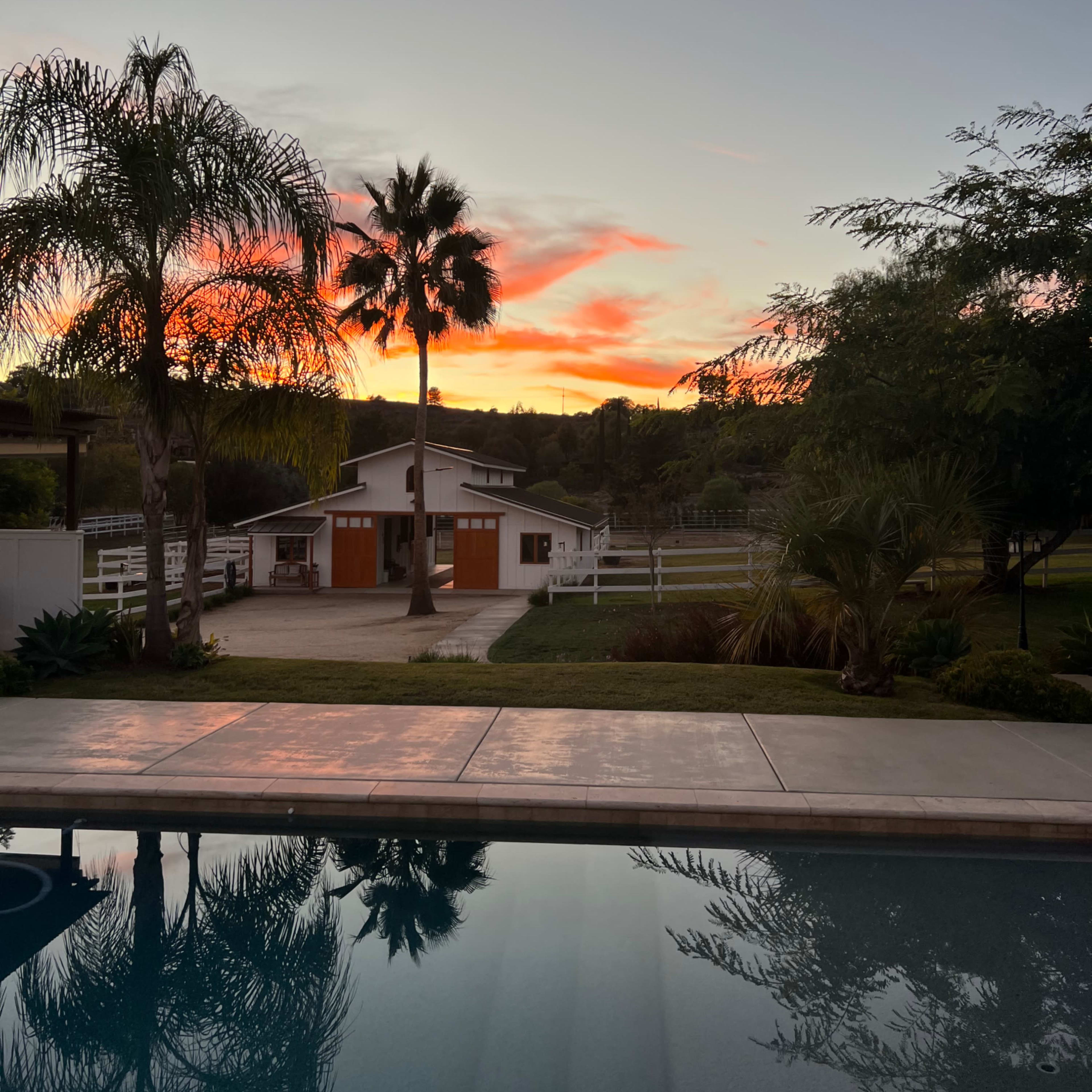 A serene view of a pool reflecting a sunset with palm trees and a barn in the background.