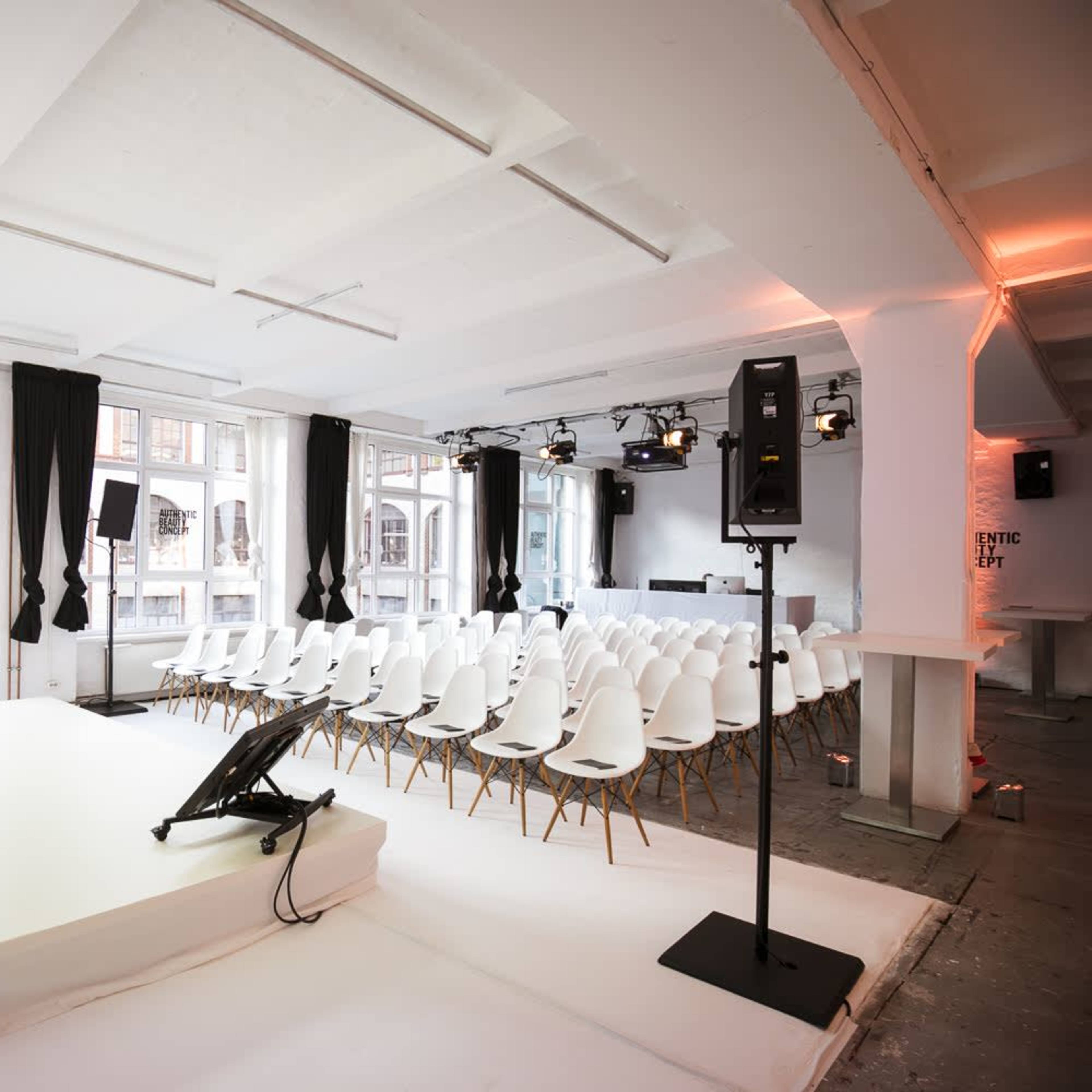 A modern event space with a white stage, rows of white chairs, and large windows allowing natural light.