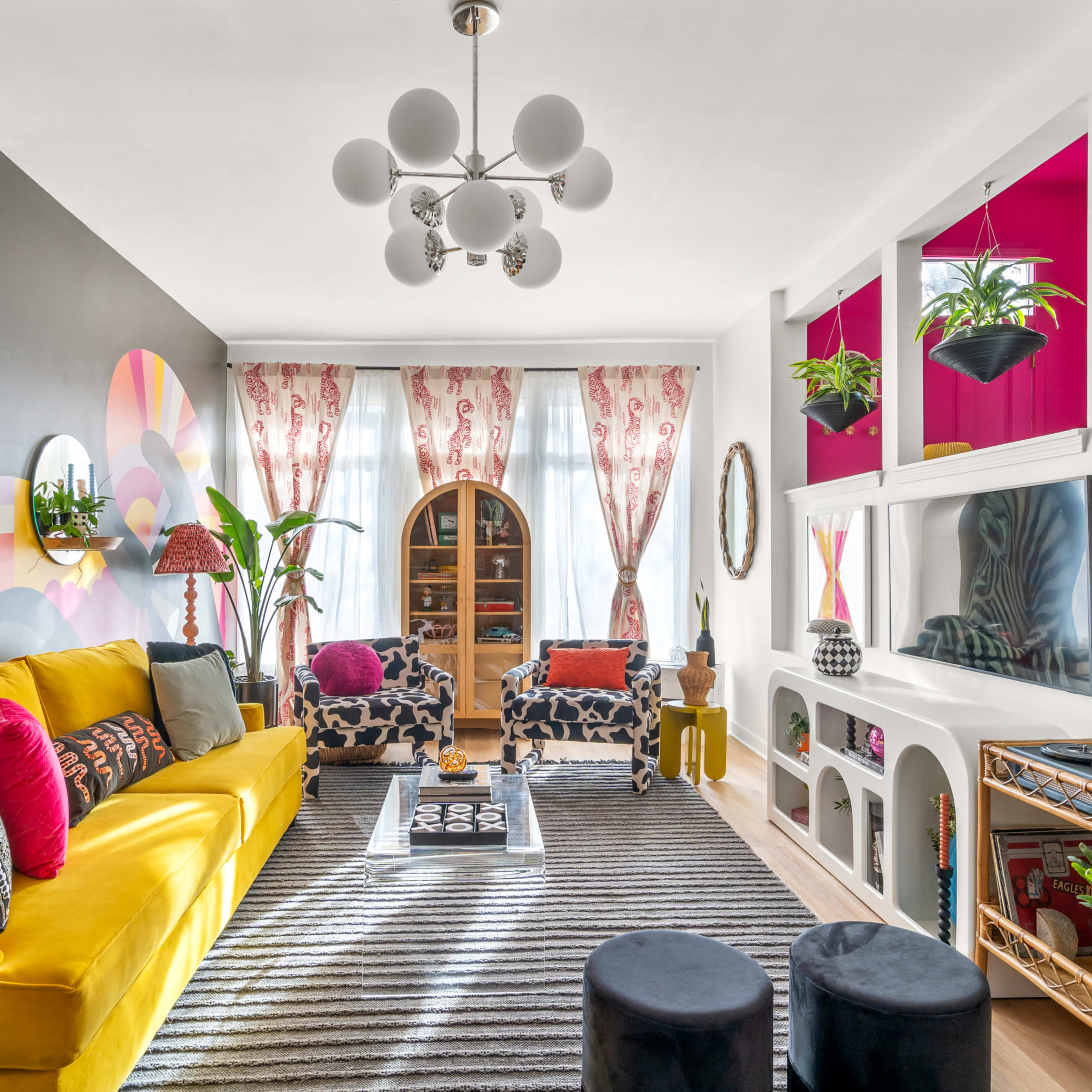 The living room features a bright yellow couch, patterned chairs, and a large mural on one wall, complemented by decorative plants and a striped rug.