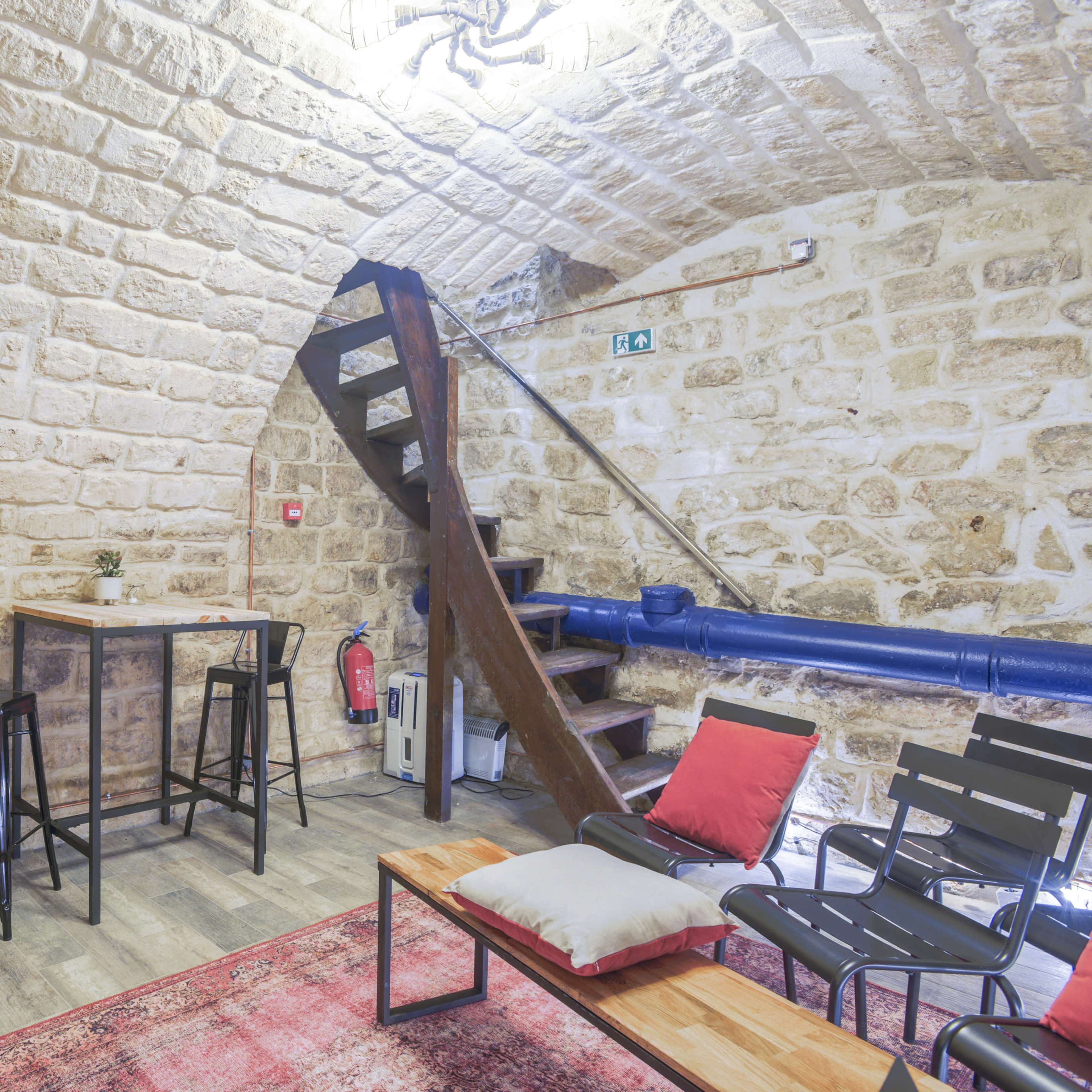 A stone-walled room featuring a small bar table with stools, a staircase, and seating areas with cushions and a rug.