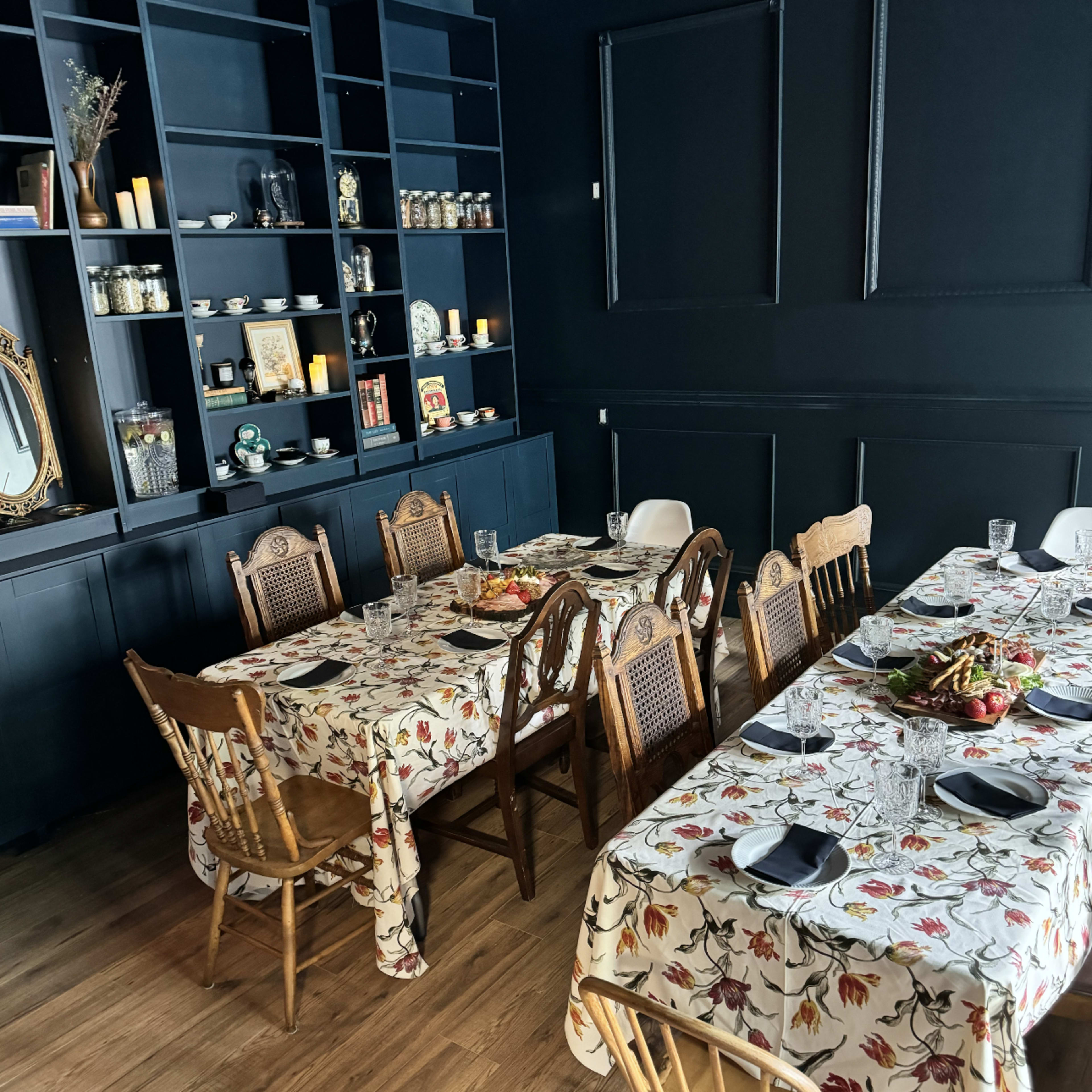 A dining area features two long tables with floral tablecloths and wooden chairs, set against a black wall lined with shelves displaying various decor items.