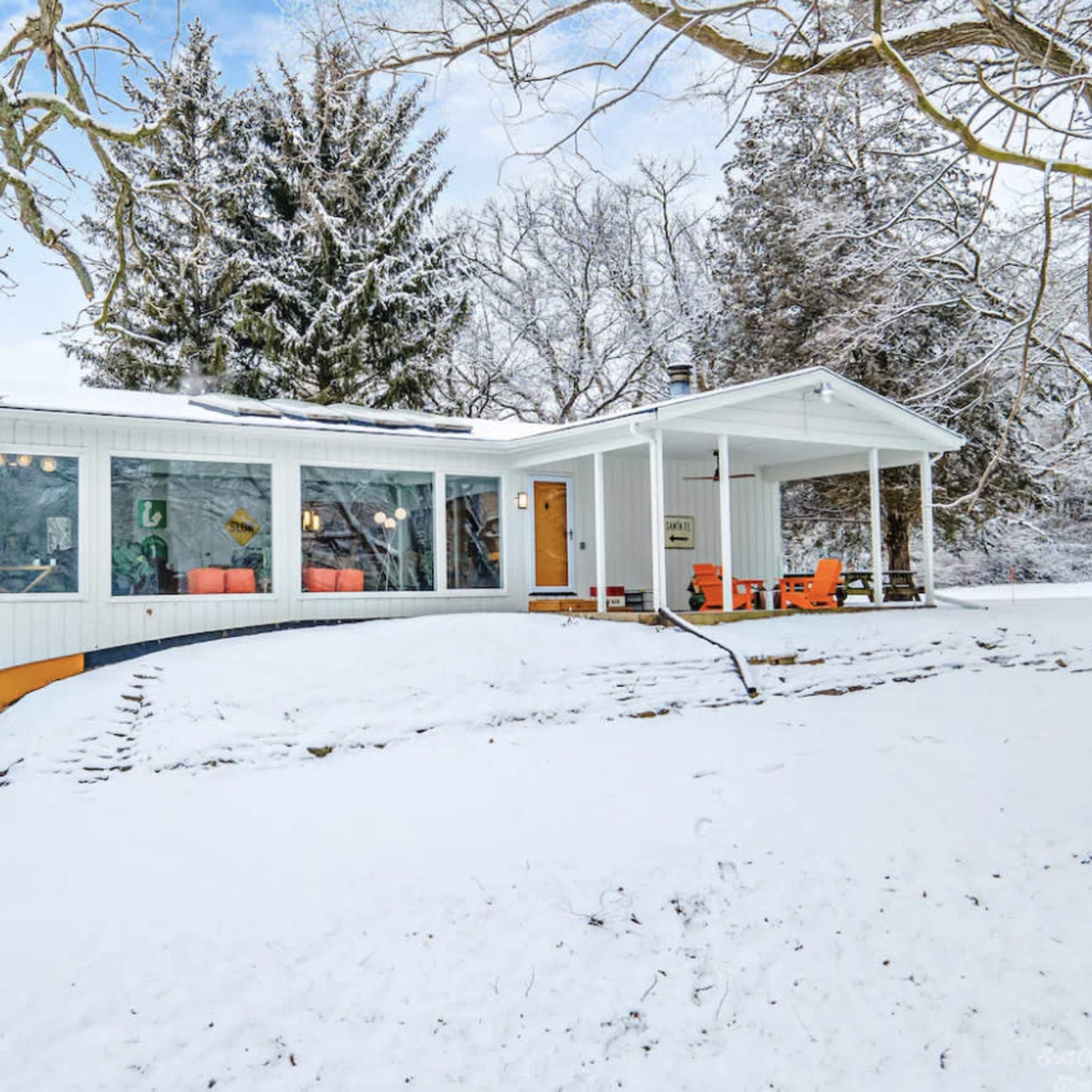 A modern house with large windows and a covered porch is surrounded by snow-covered ground and trees.