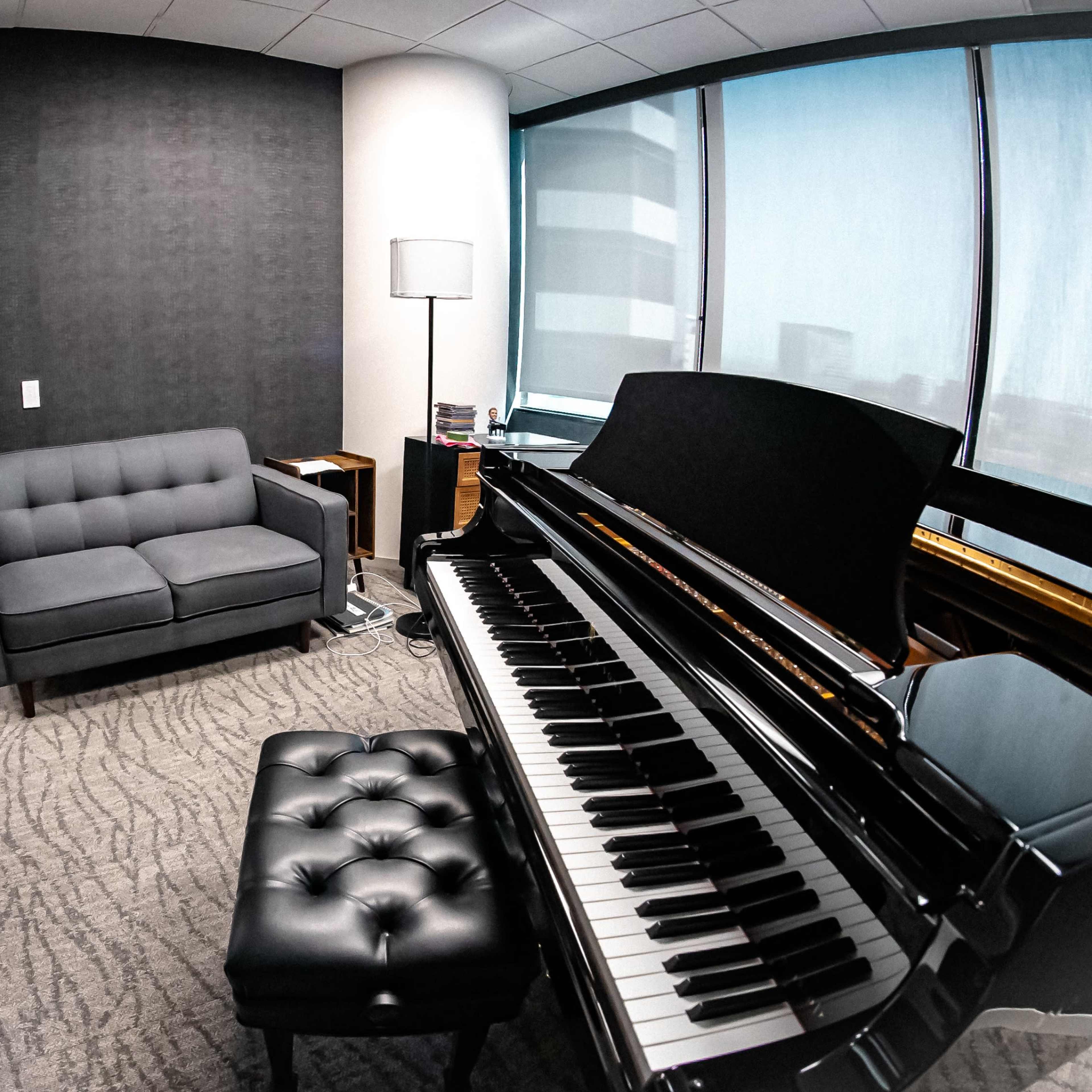 A corner of an office space featuring a black piano, a gray couch, a lamp, and large windows.
