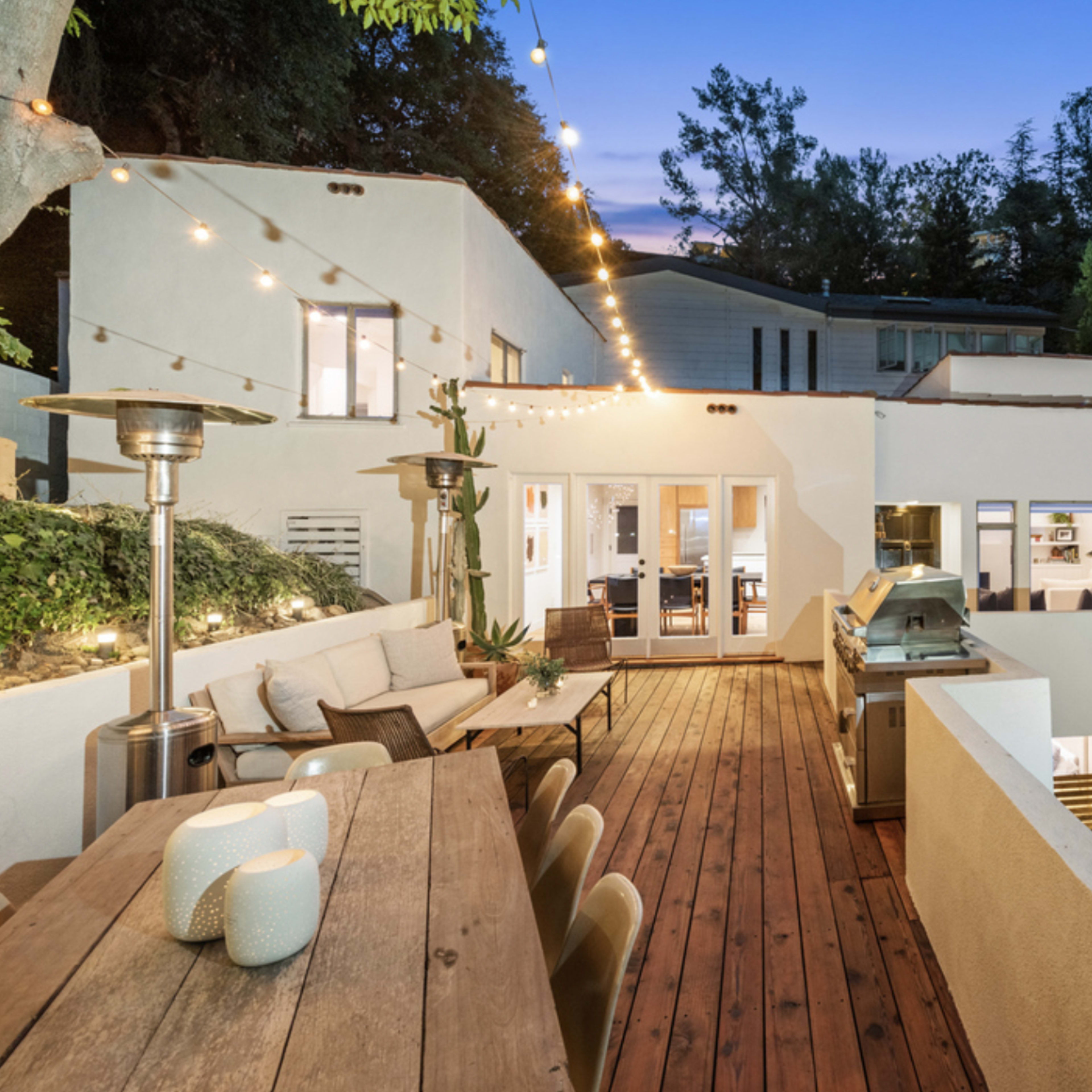 A modern home features a spacious deck with a dining area, outdoor heaters, and string lights, surrounded by greenery and a barbecue area.
