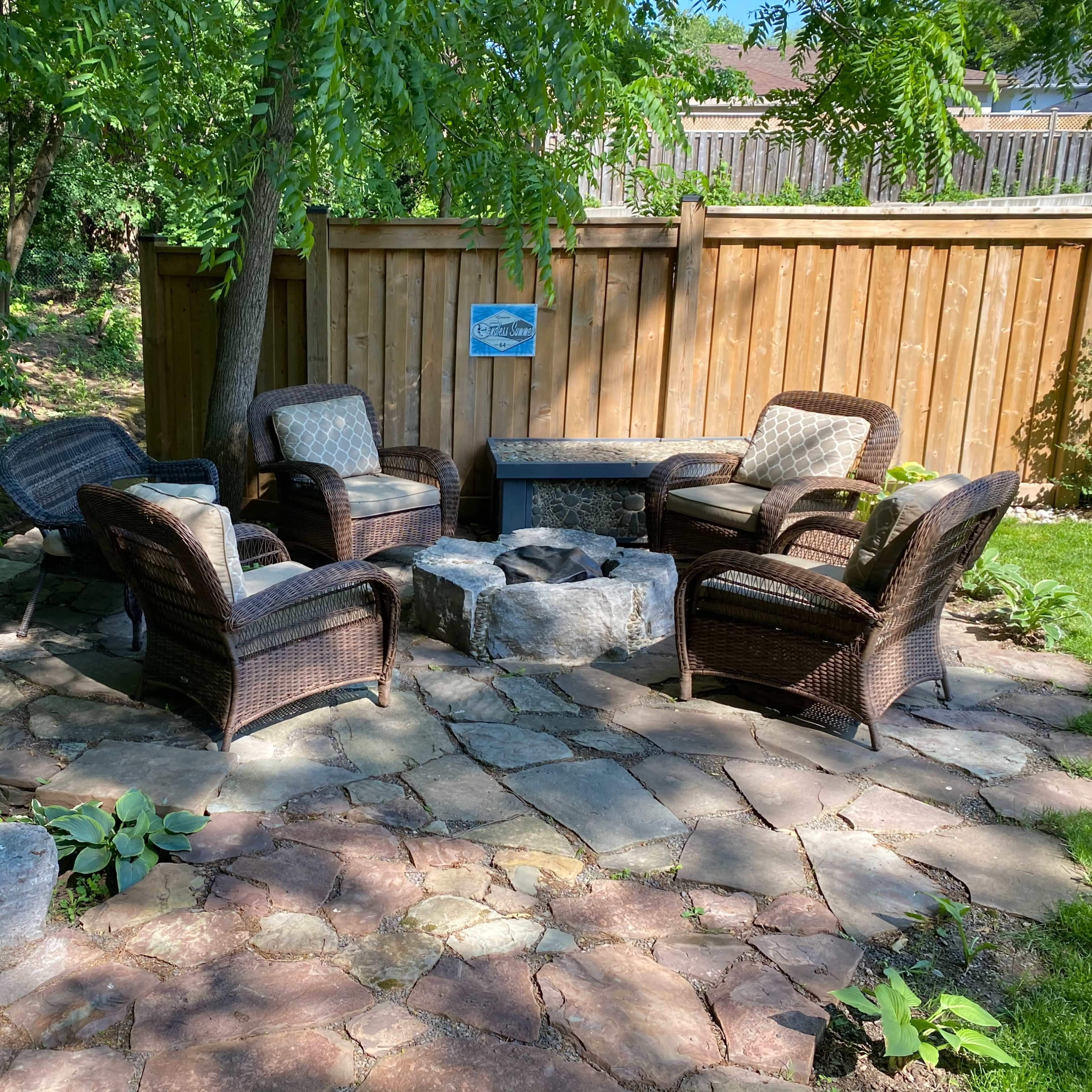 A seating area with four wicker chairs arranged around a circular stone fire pit is set within a landscaped yard surrounded by trees and a wooden fence.