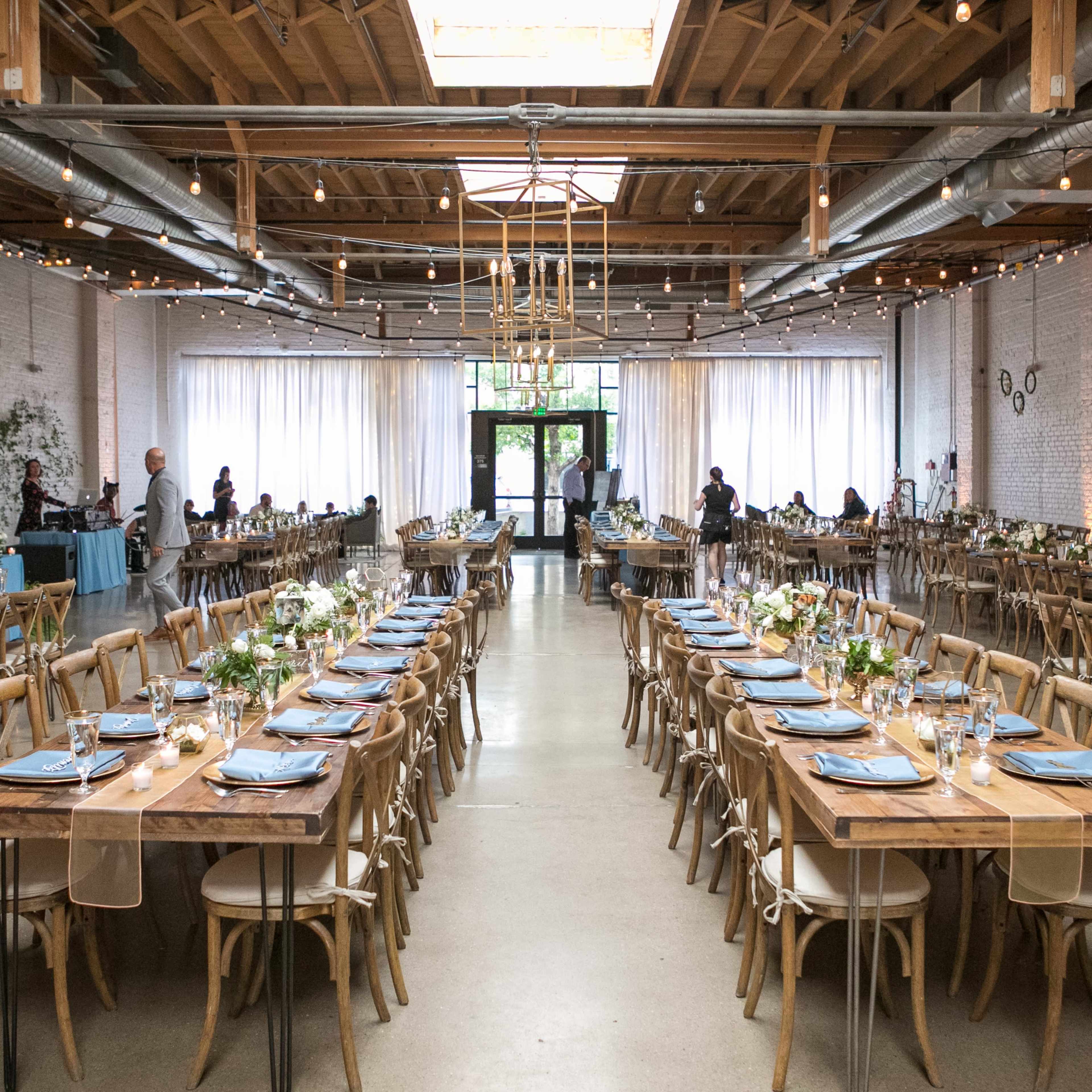 A spacious, elegantly arranged dining hall features long wooden tables set with blue napkins and floral centerpieces, illuminated by hanging lights and natural daylight.