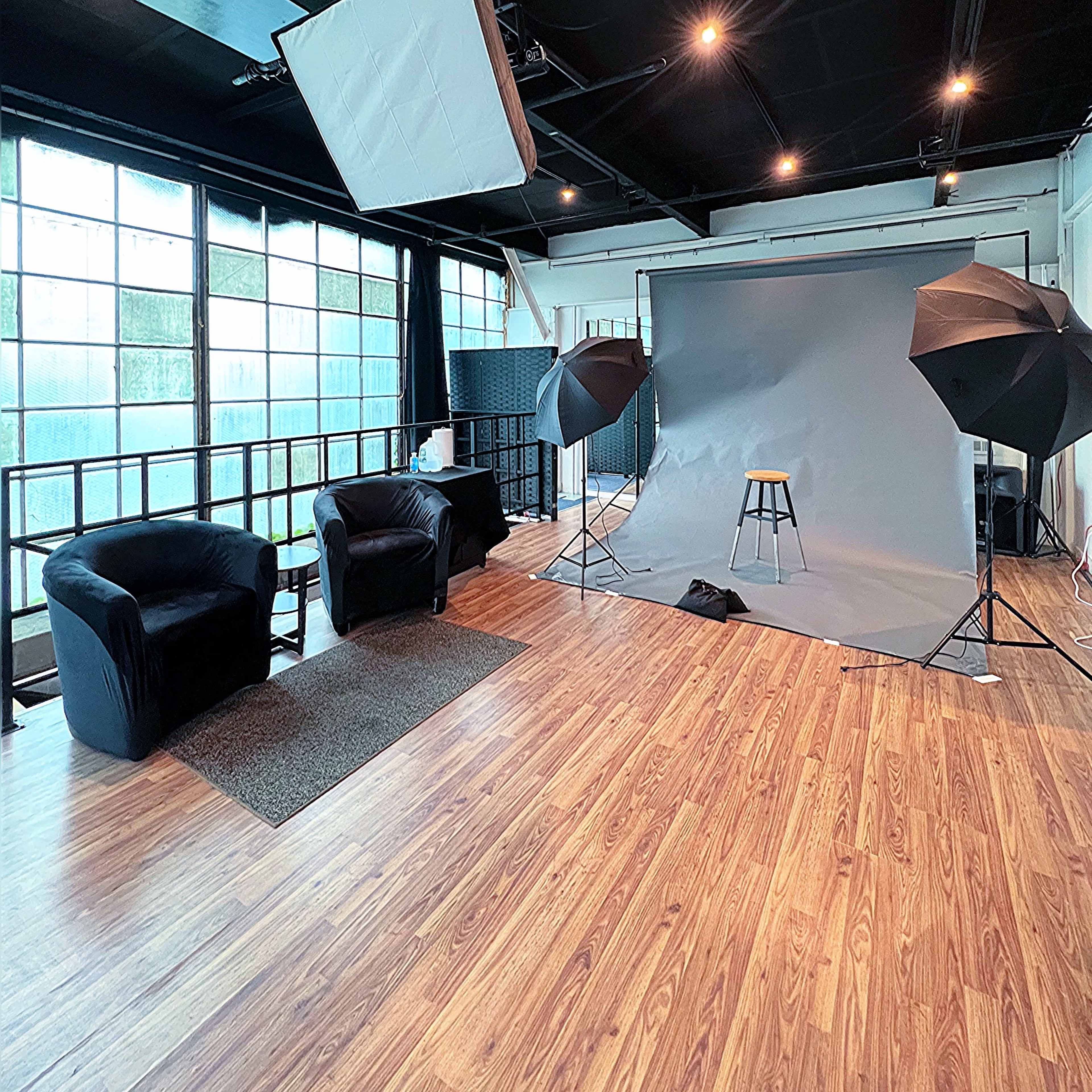 A photography studio features a gray backdrop, a stool, two softbox lights, and two black chairs on a wooden floor.