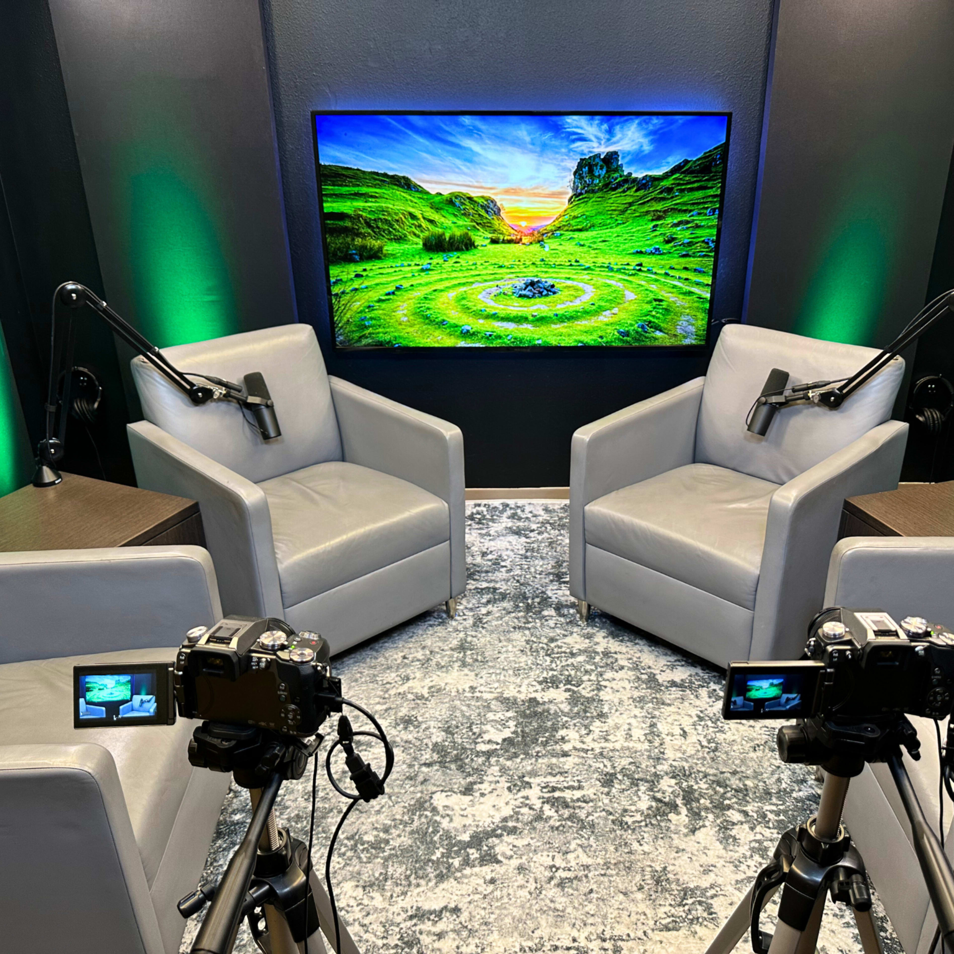 A modern interview setup with two gray armchairs facing a large screen displaying a scenic landscape, while two cameras are positioned on tripods in front of the chairs.