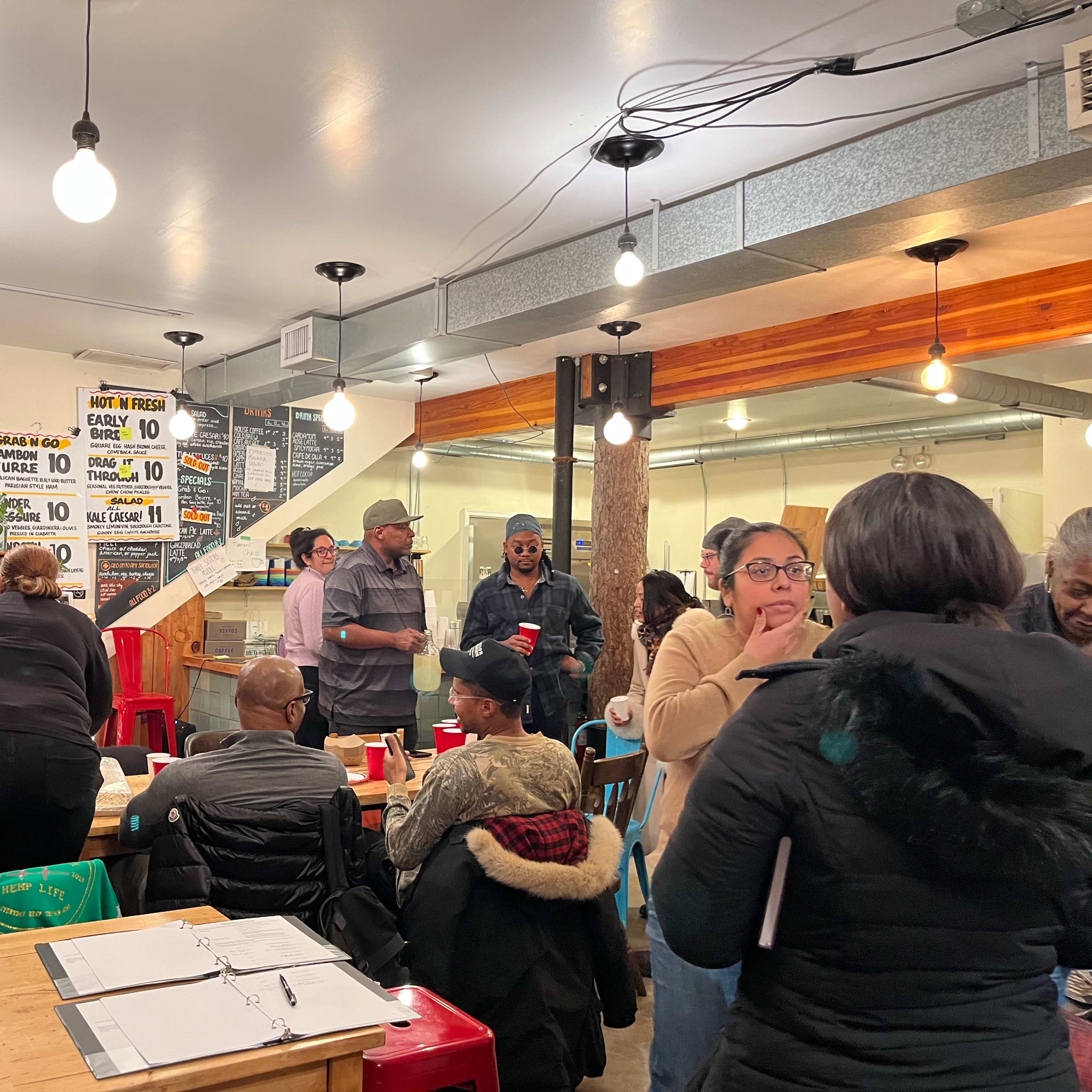 A busy restaurant interior features people dining and socializing around tables while a menu board is visible in the background.