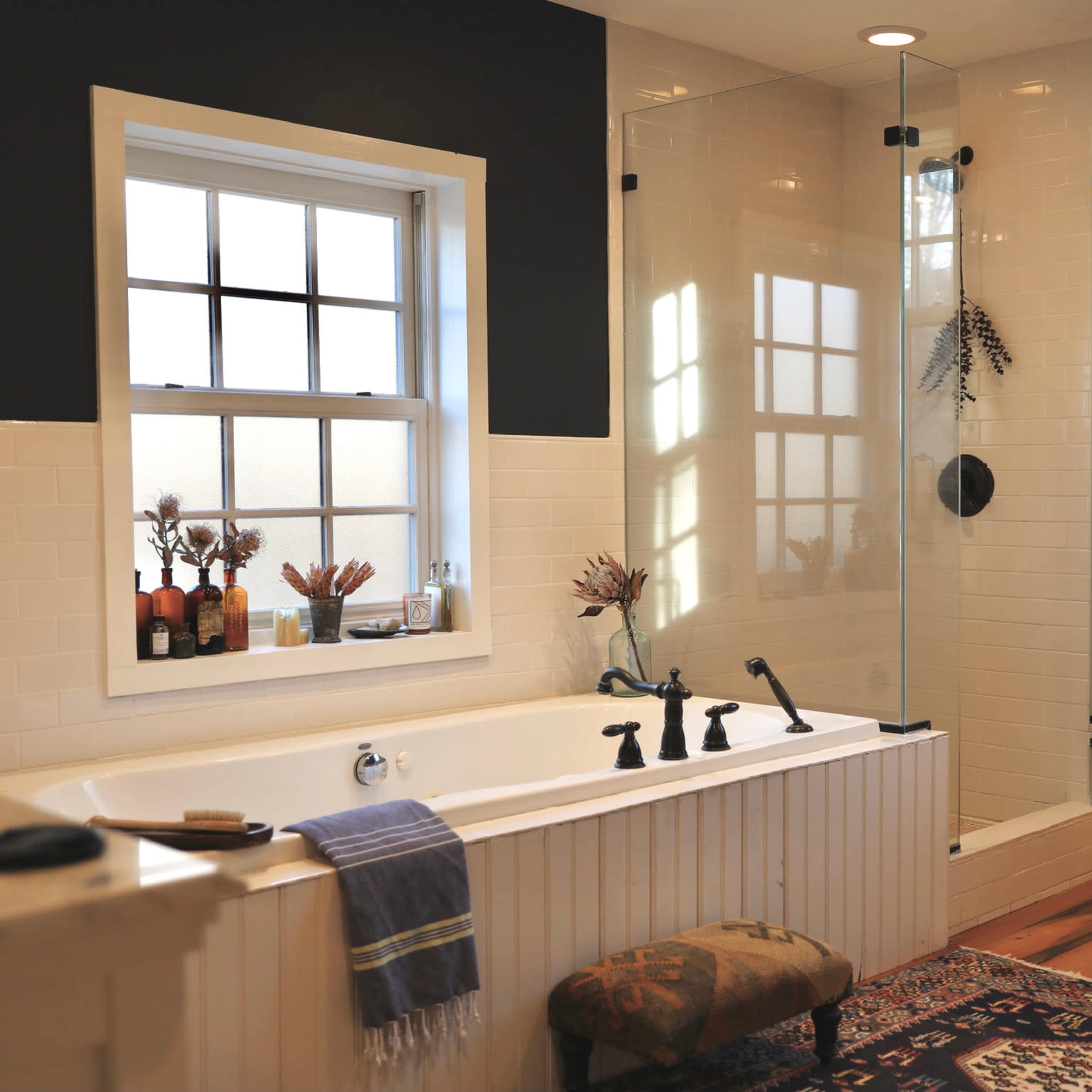 The bathroom features a freestanding tub, a glass-enclosed shower, and a large window, with wood flooring and a decorative rug.