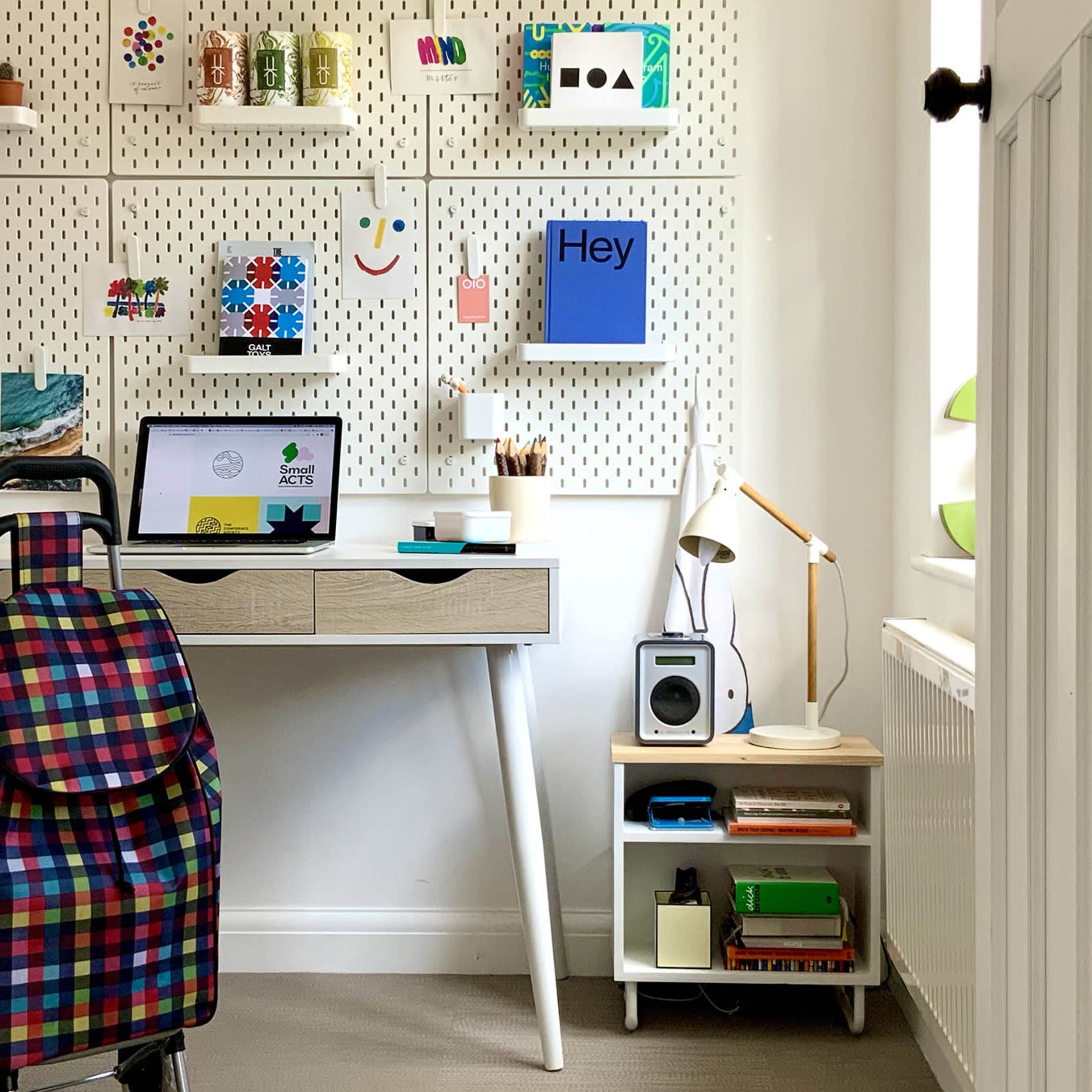 A modern workspace features a wall with colorful decorations and a desk with a laptop, a lamp, and organized supplies beside a shelf with books and a speaker.