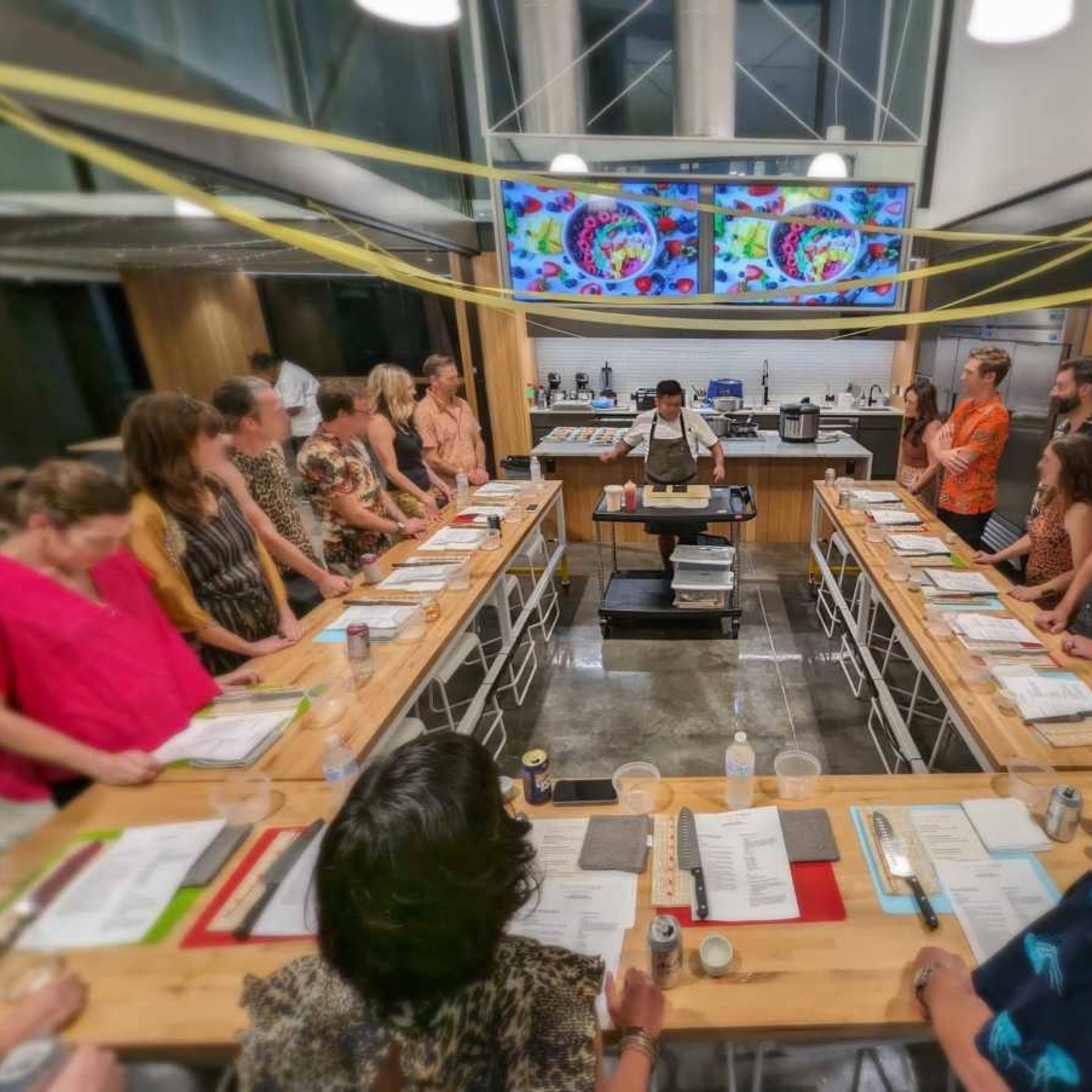 A group of people gathers around a long wooden table in a modern kitchen, attentively watching a chef demonstrating cooking techniques.