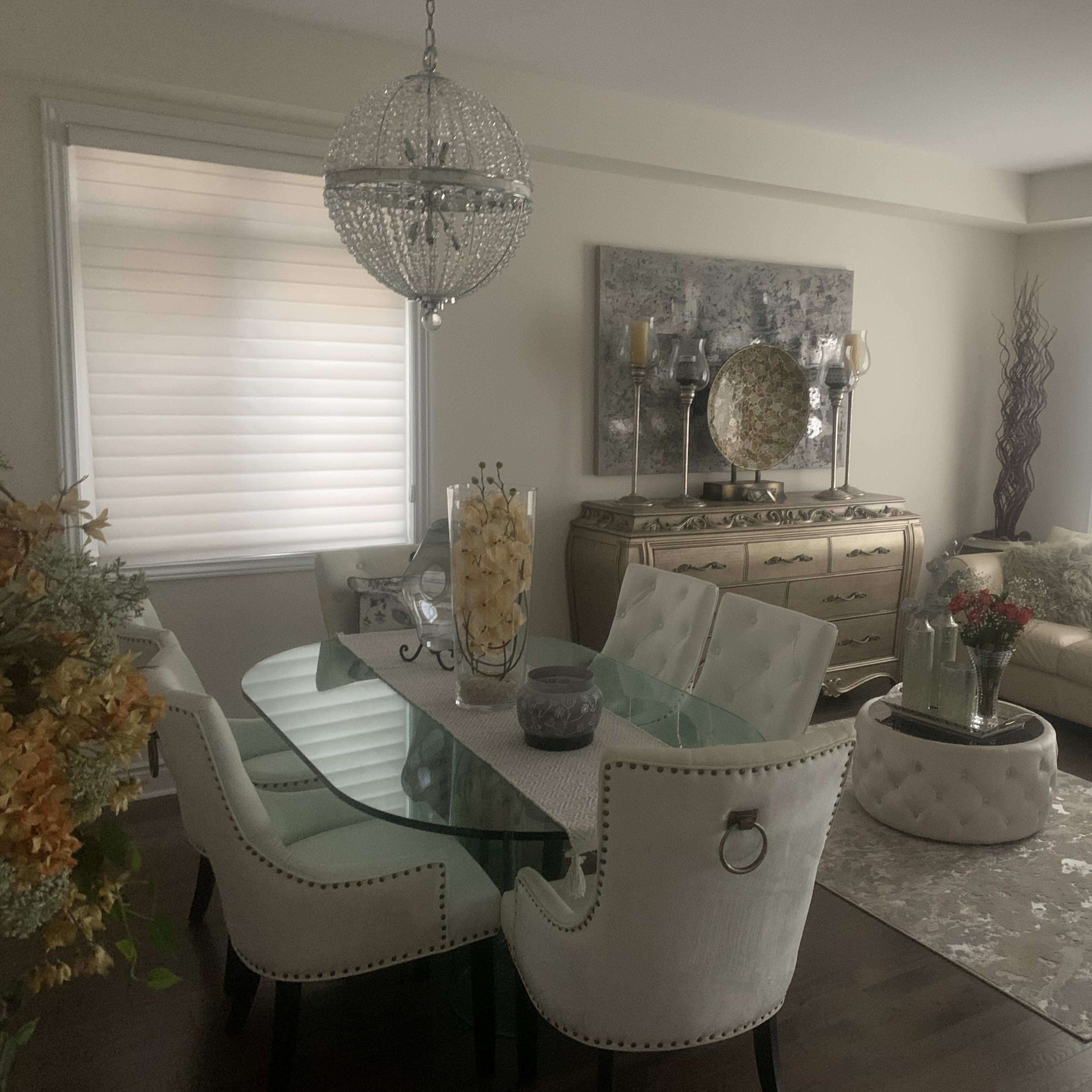 A modern living and dining area features a glass dining table surrounded by upholstered chairs, a light-colored sofa, and decorative elements along with a large wall art piece.