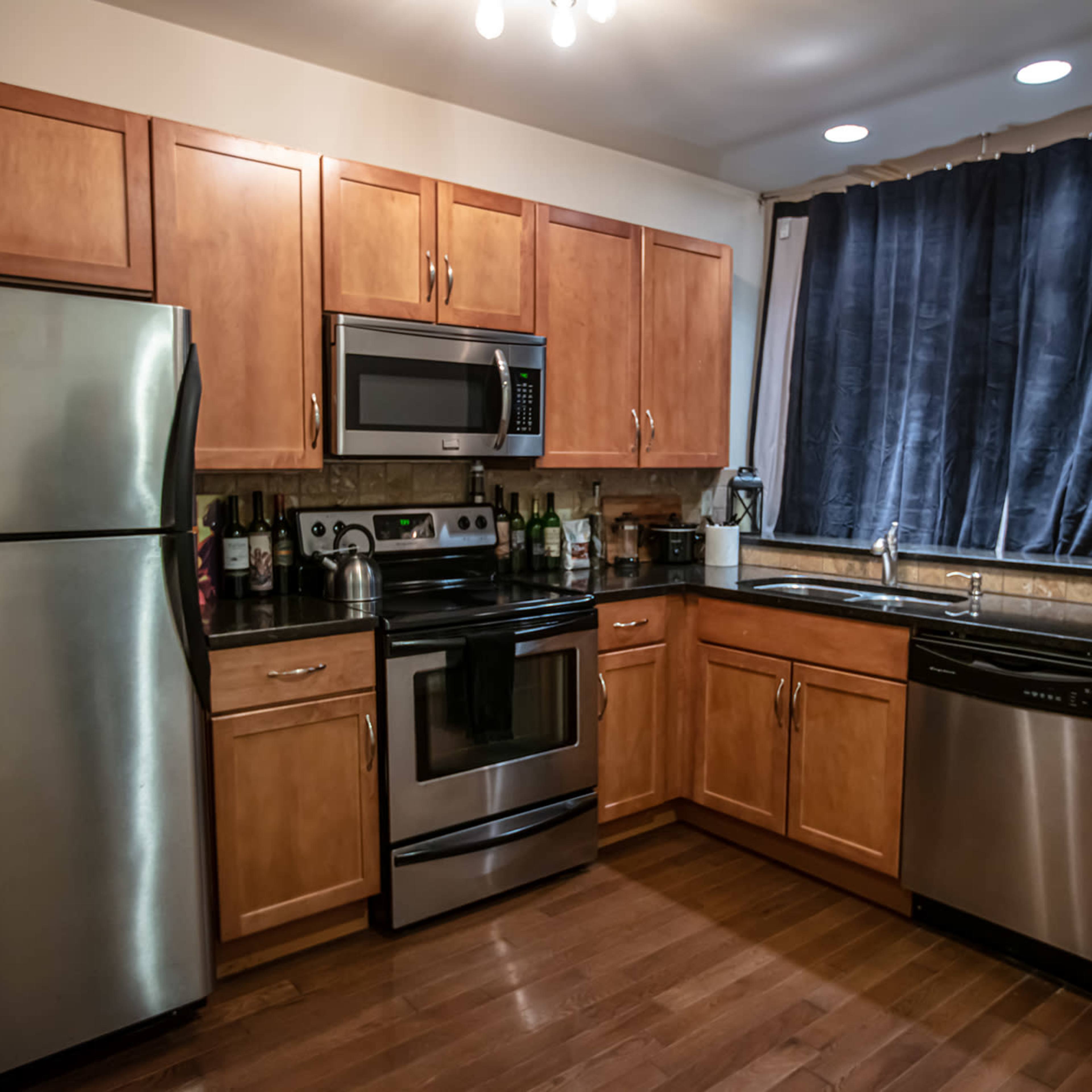 A kitchen equipped with wooden cabinets, a stainless steel refrigerator, an oven, a microwave, and a dishwasher, with dark curtains on the window.