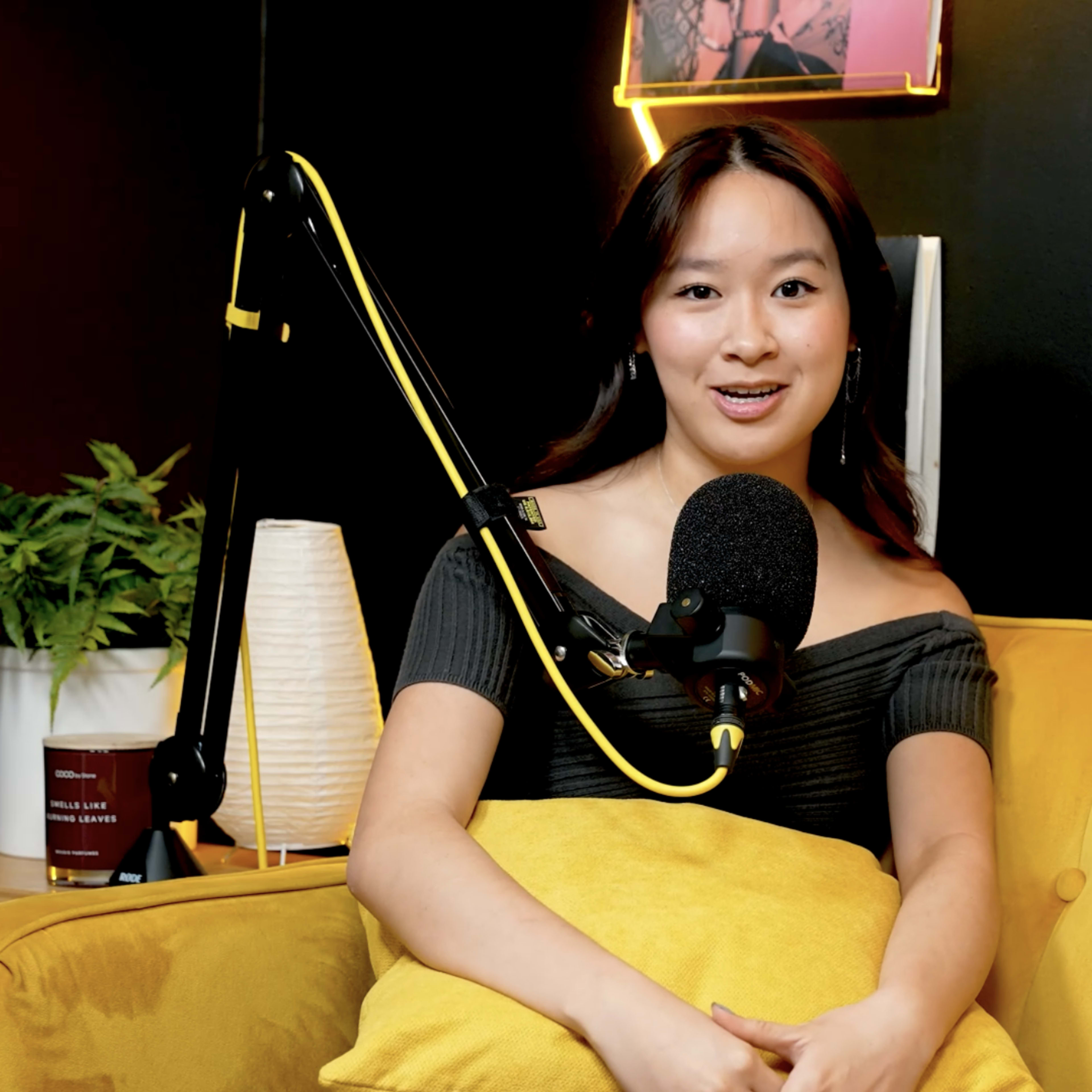 A person sits on a yellow couch, holding a yellow blanket, with a microphone setup in front of them and framed artwork on the wall behind.